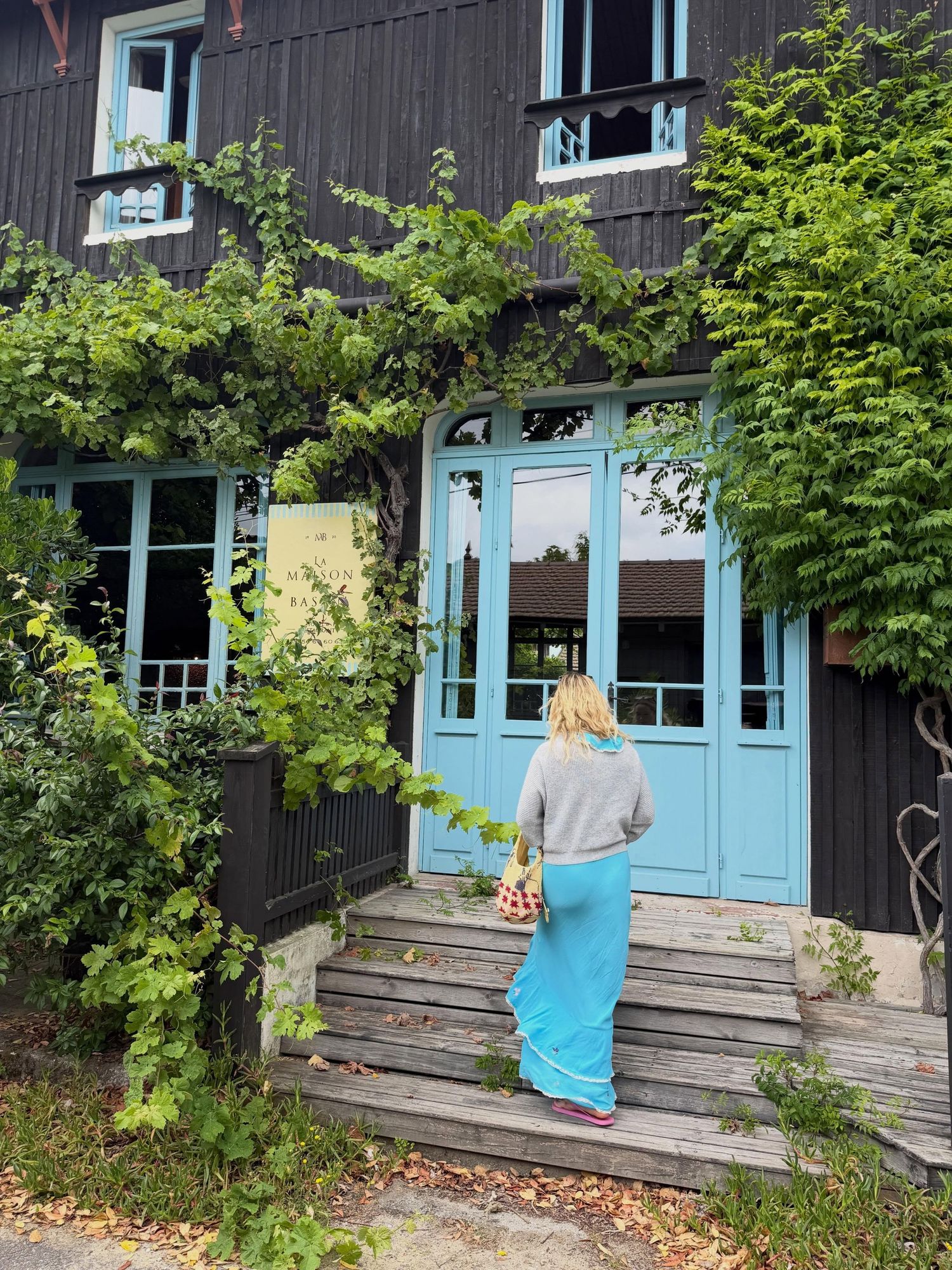 Woman in blue skirt approaching a dark wooden building with turquoise doors and lush green ivy.
