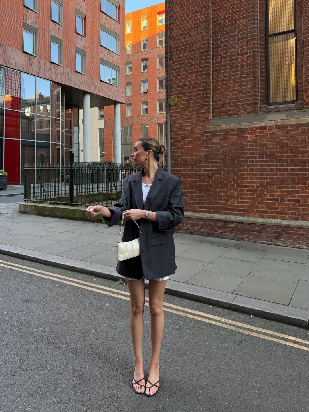 Woman in oversized blazer, mini shorts, and strappy heels holding white bag on urban street.