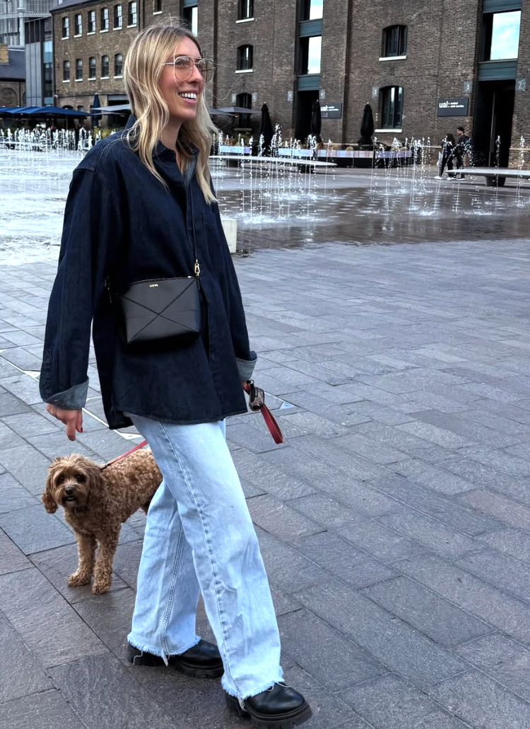 A person in a black top and light jeans walks with a brown goldendoodle dog on a paved outdoor plaza.