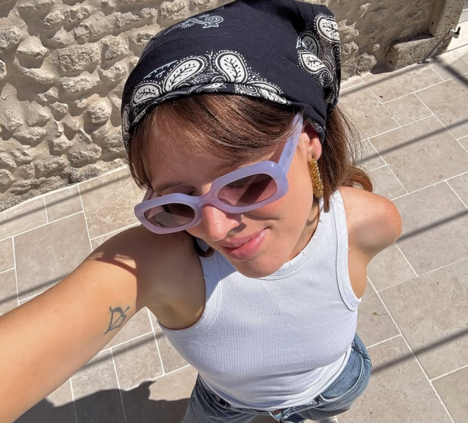 Person in a baseball cap and sunglasses taking a selfie against a stone wall backdrop.