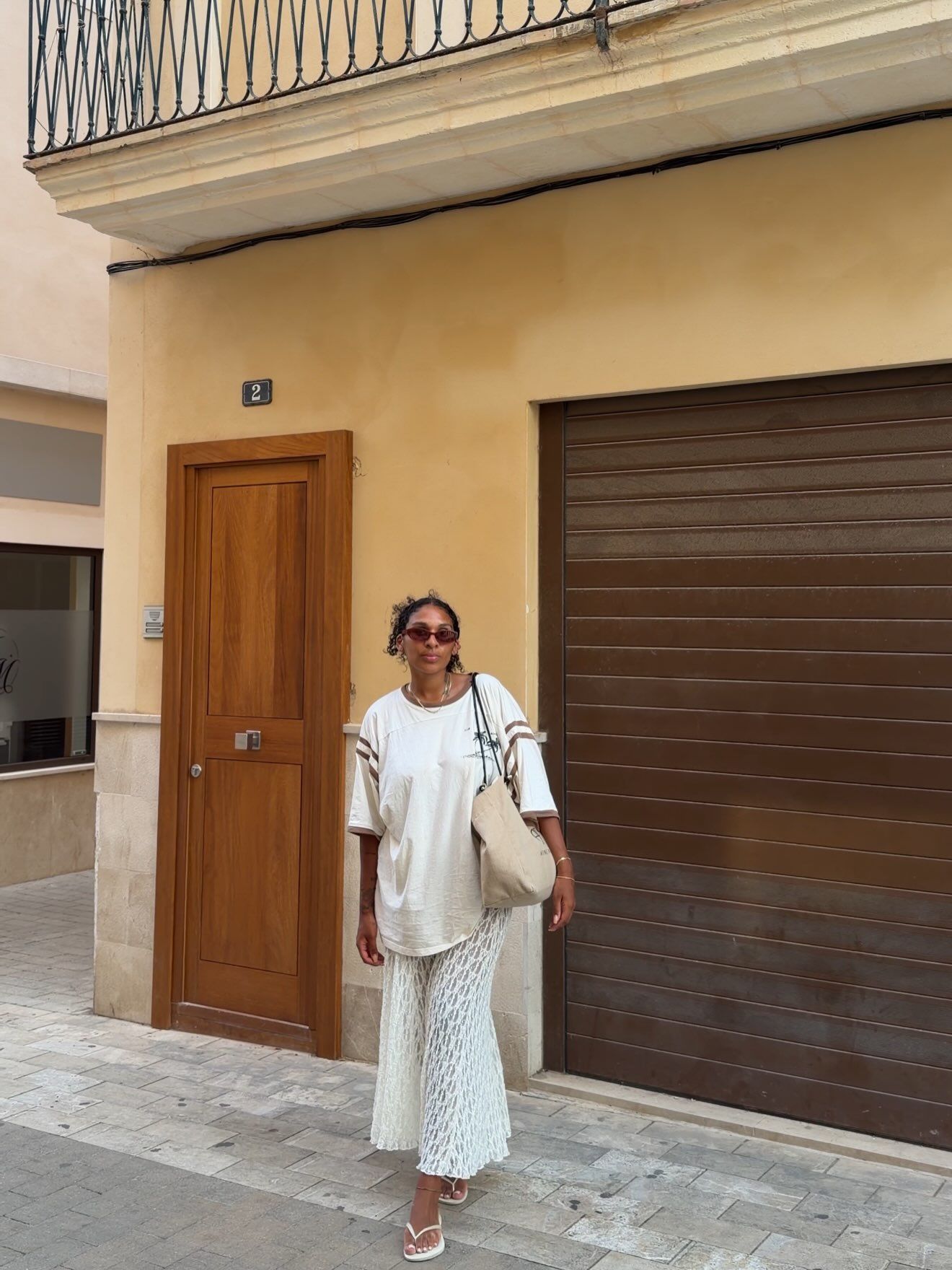 Woman in white oversized outfit and sunglasses standing outside Mediterranean building.