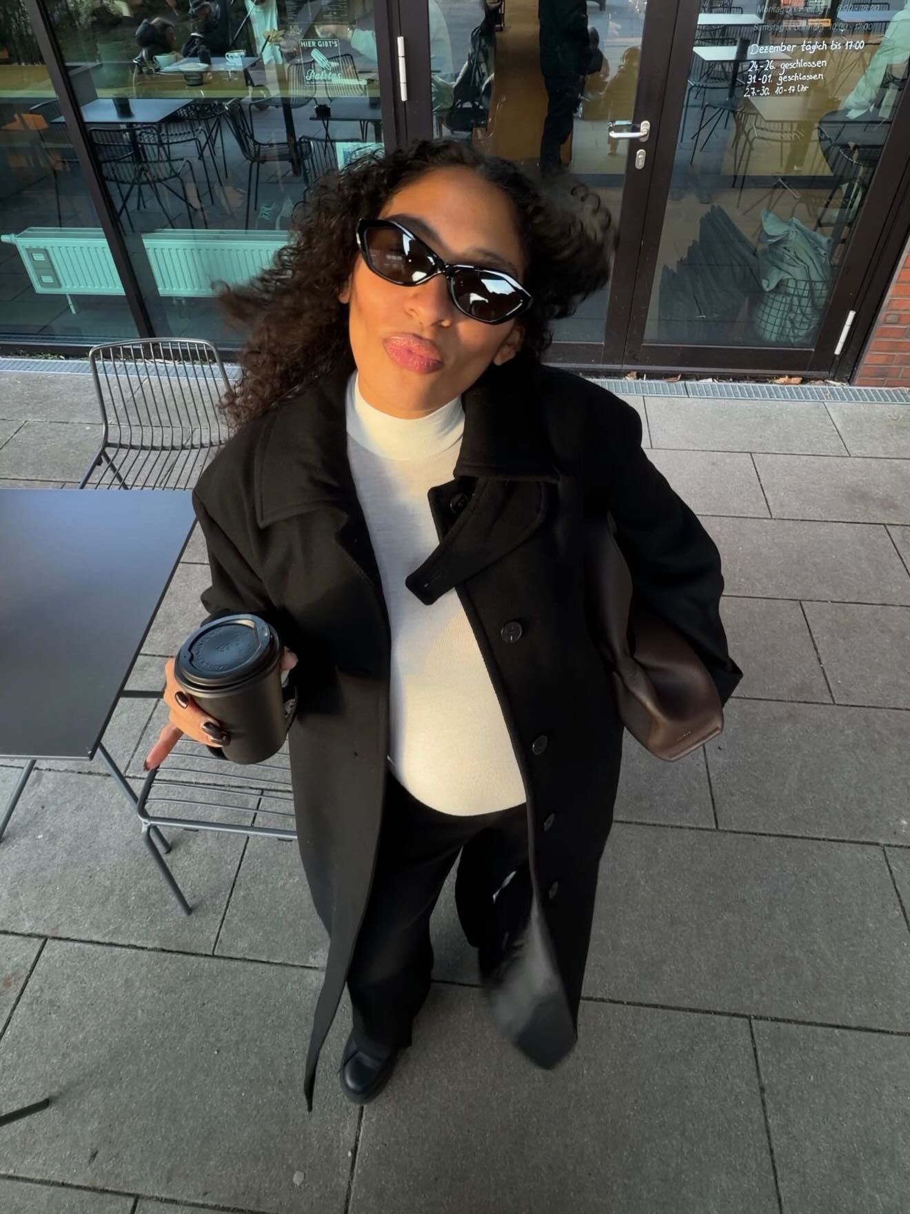 Woman in black trench coat and sunglasses holding coffee cup outdoors.