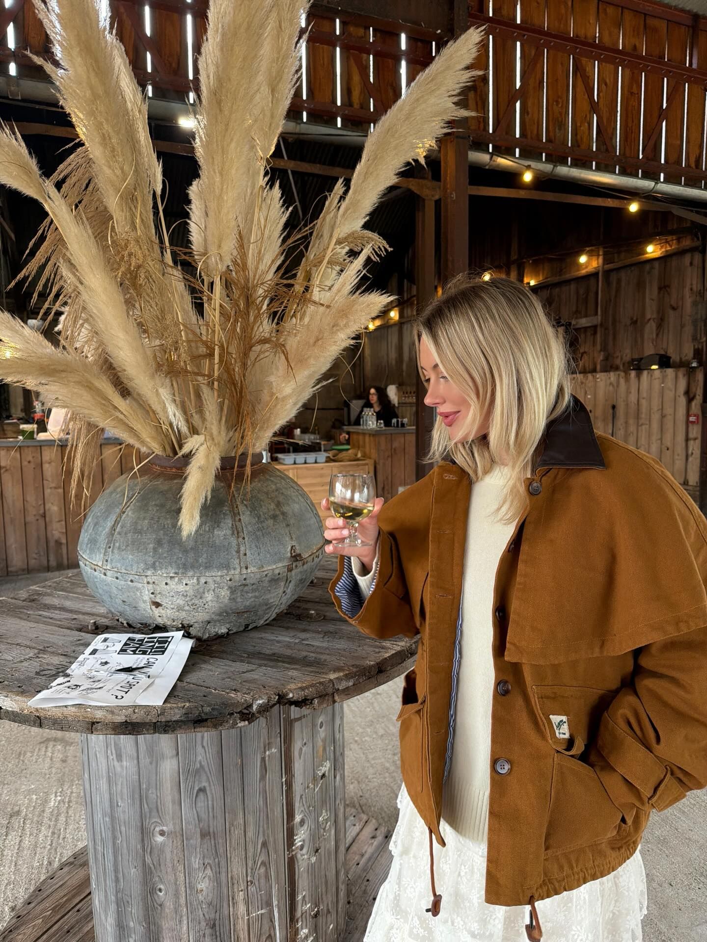 Woman in caramel jacket holding white wine glass beside pampas grass arrangement in rustic barn setting.