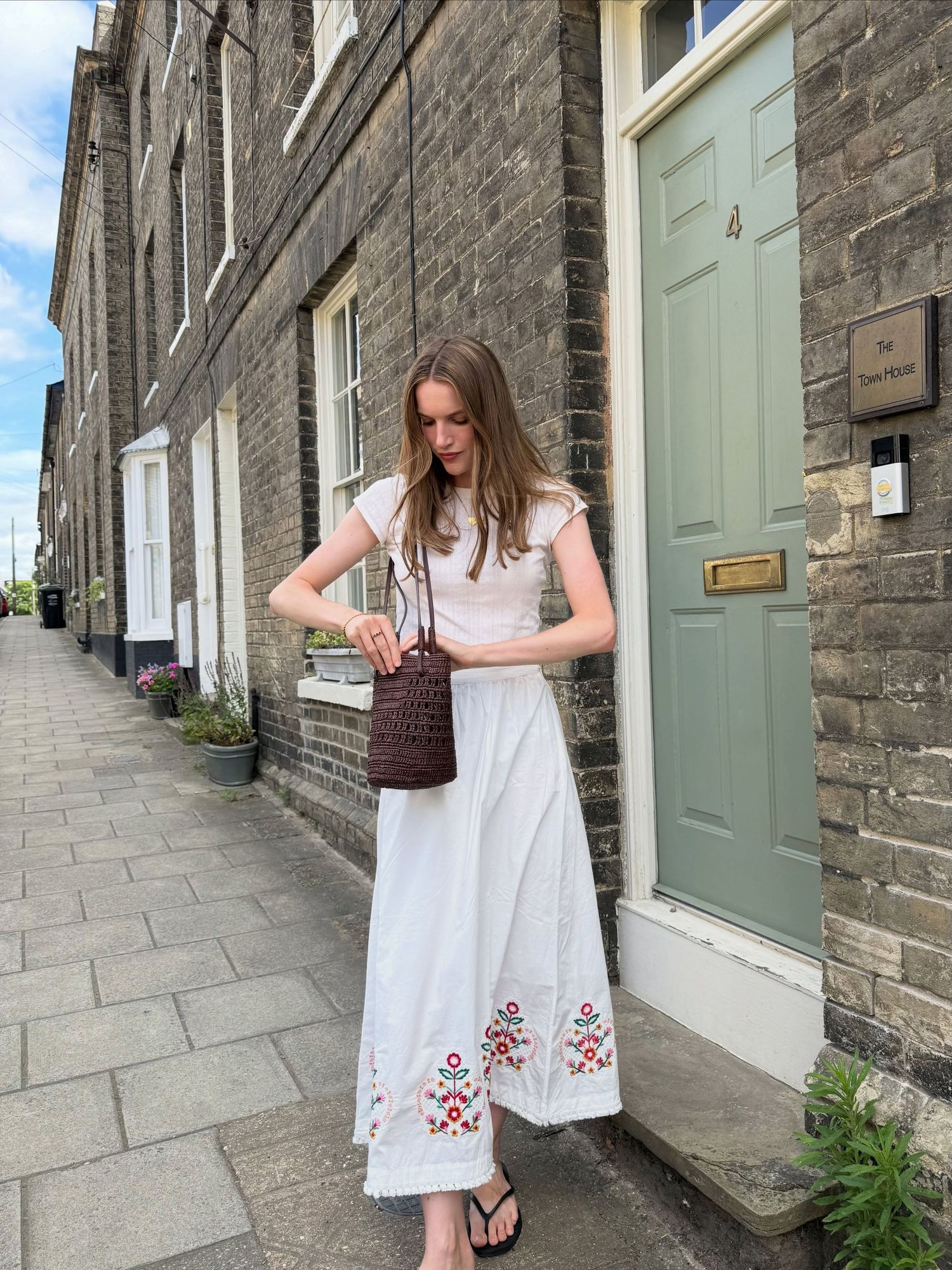 Woman in white embroidered maxi skirt and white top holding a brown wicker bag on a cobblestone street.