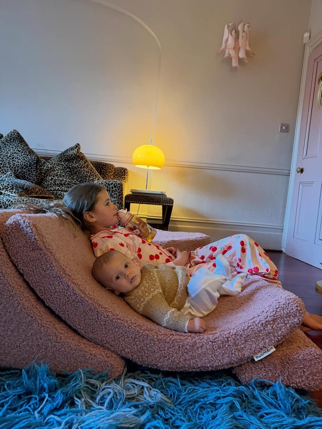 Two young children relaxing together on a pink fluffy bean bag chair in a cozy room with warm lamp lighting.