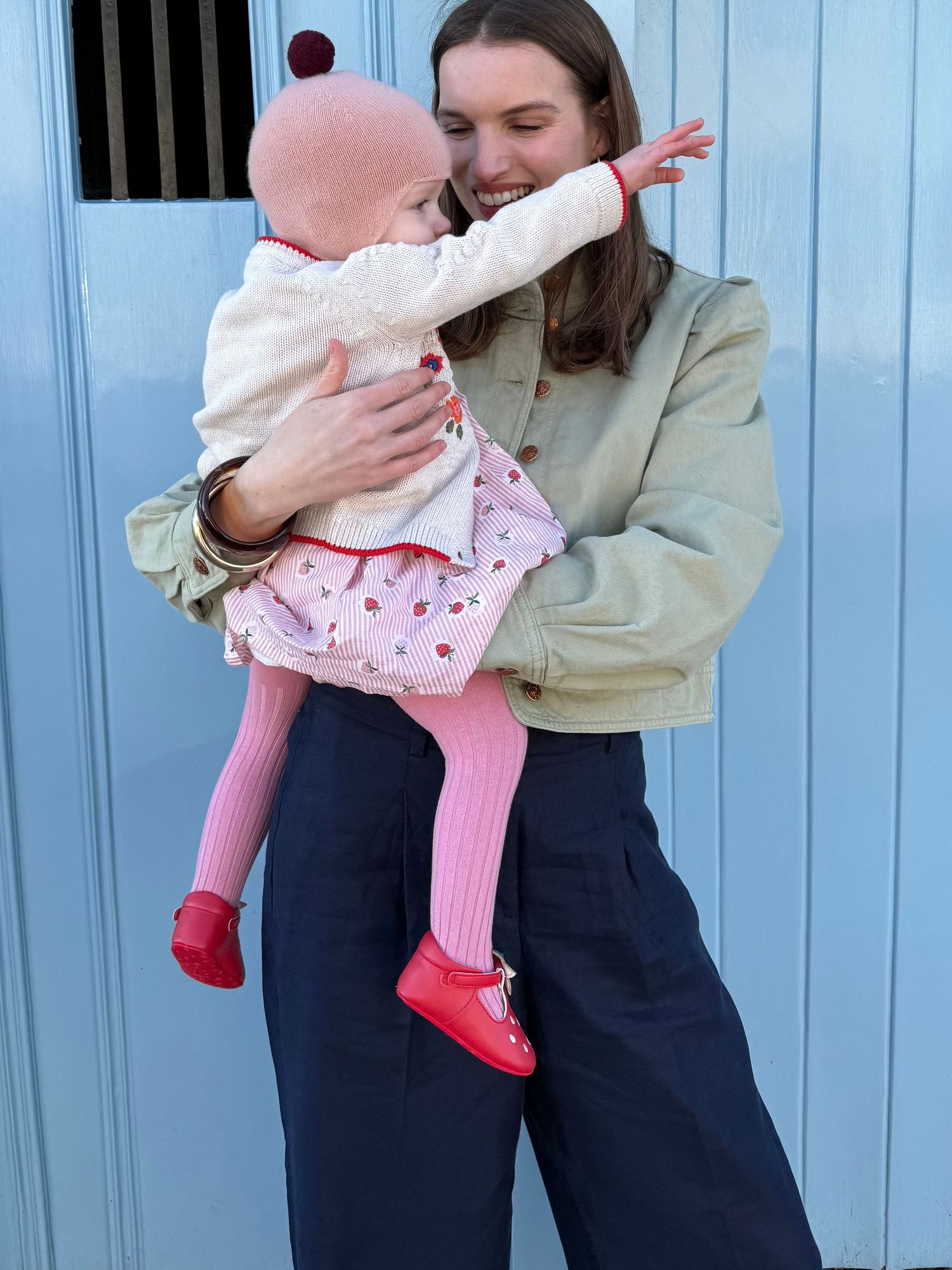 A baby in pink clothing and red shoes being held while reaching out against a blue door background.