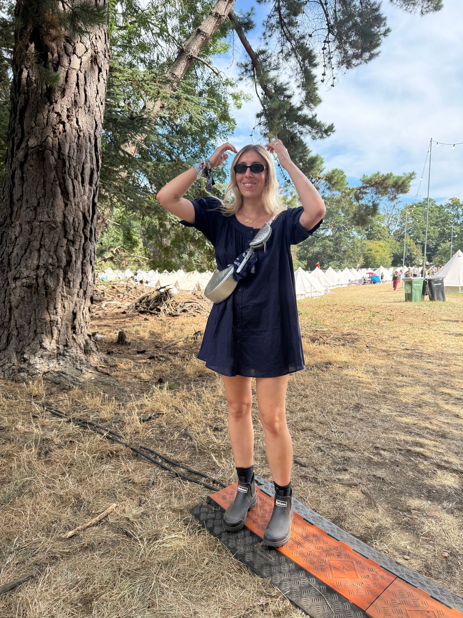 Woman in navy dress and black socks with wedge shoes stands under pine tree at outdoor festival on sunny day.