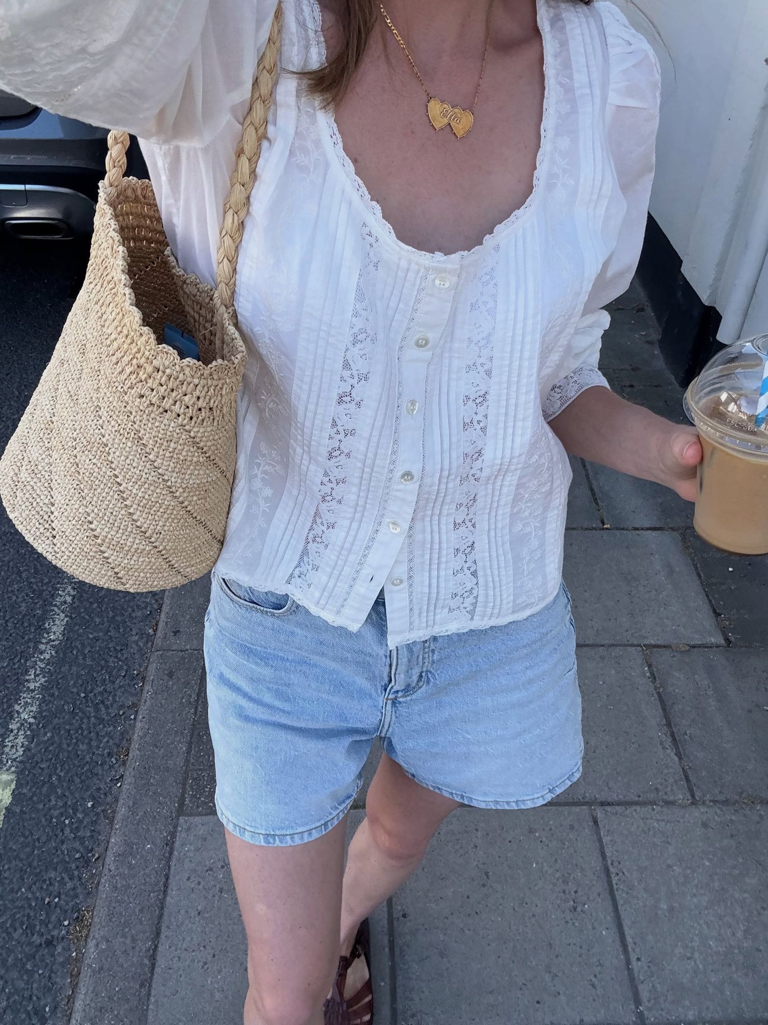 Woman in white lace blouse, denim shorts, gold heart necklace, holding iced coffee, carrying woven bag outdoors.