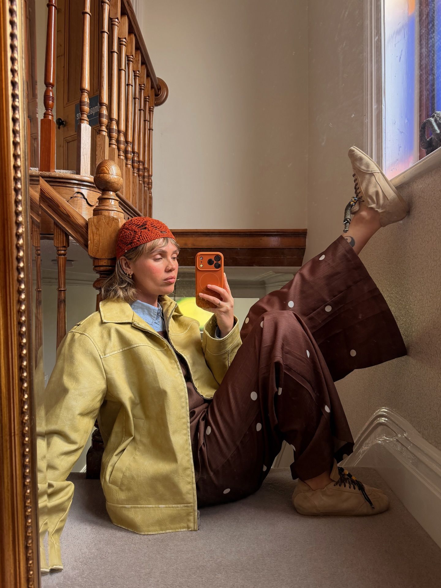Woman in yellow jacket, brown polka dot pants, and orange beanie takes mirror selfie on staircase floor.