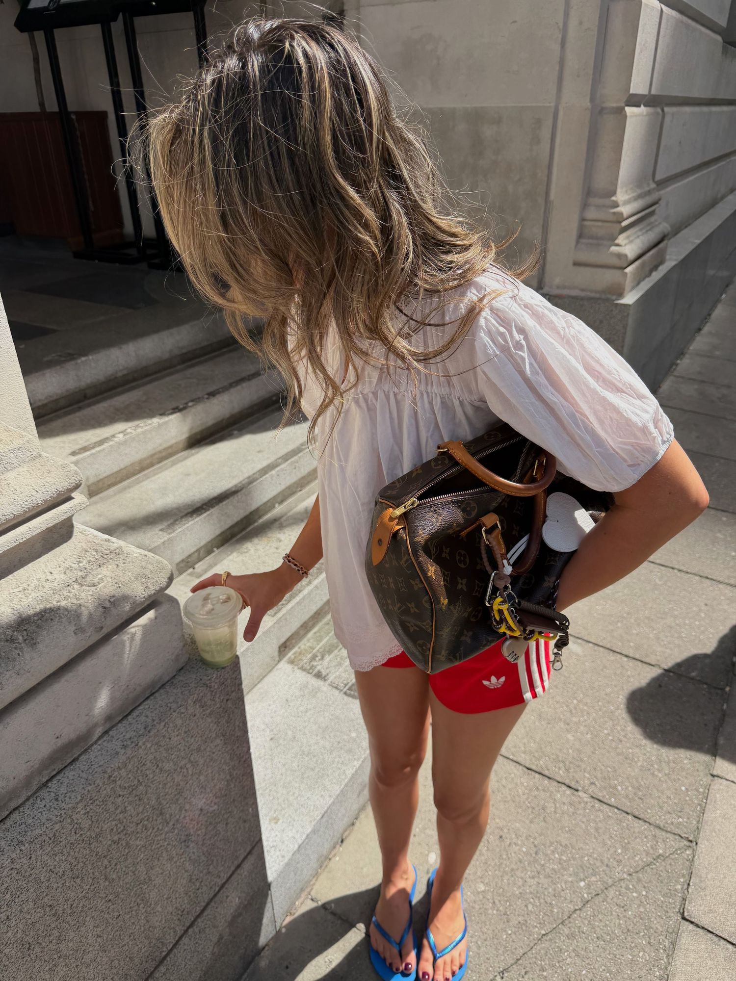 Woman in white top, red Adidas shorts, blue flip flops, carrying Louis Vuitton bag on sunny city steps.