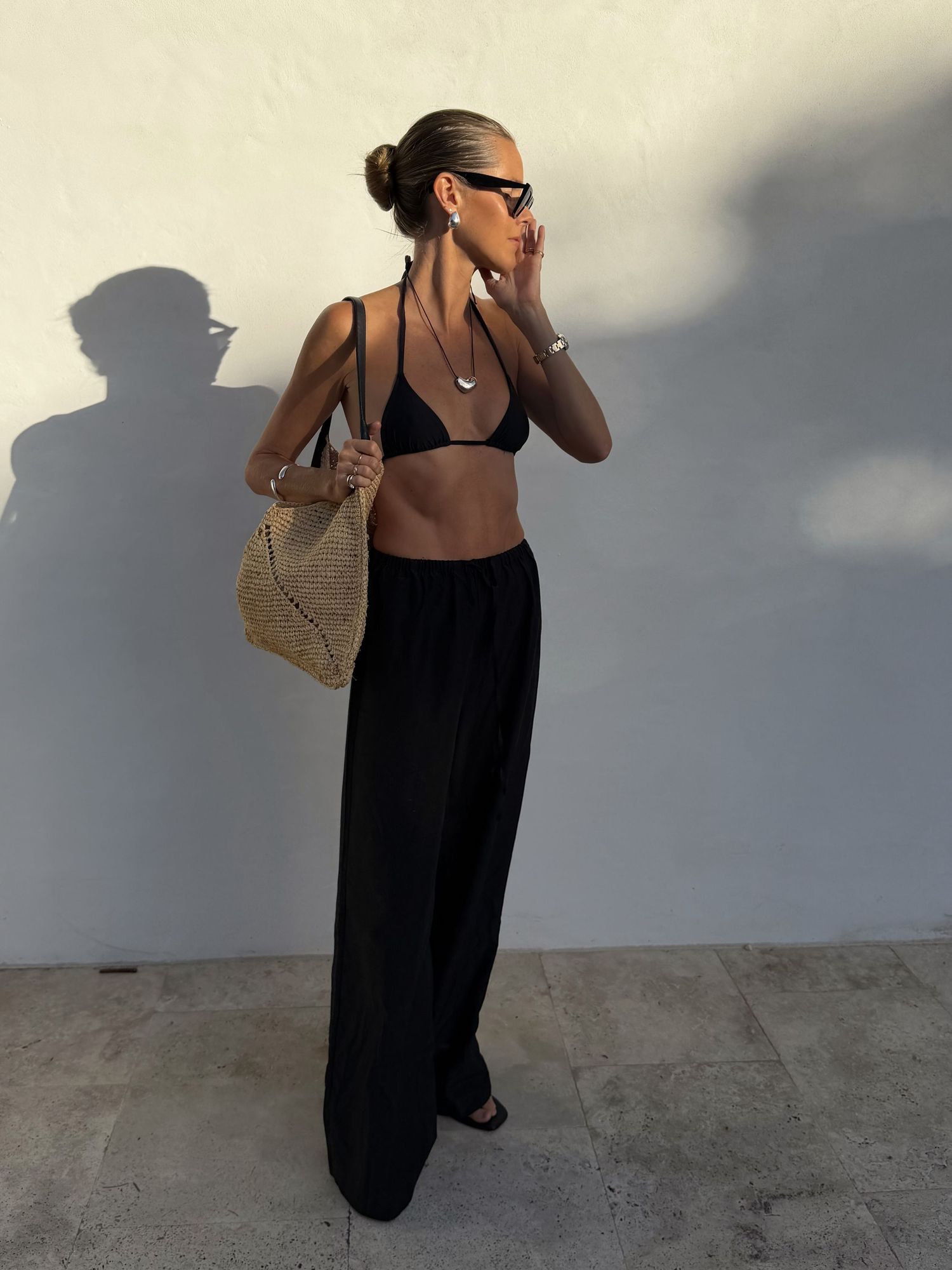 Woman in black bikini top and wide-leg pants with woven bag and sunglasses posing against white wall.