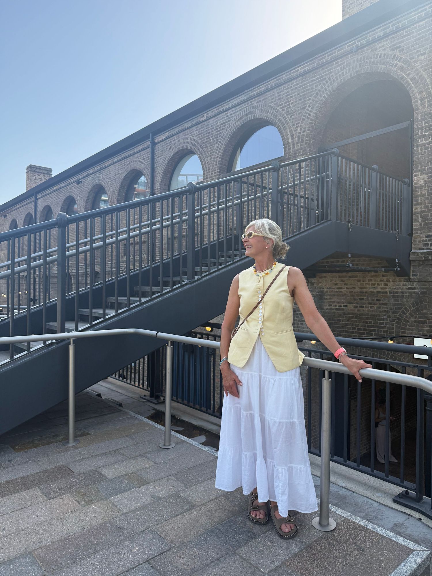 Woman in yellow top and white skirt stands by metal railing near brick building with arched windows on sunny day.