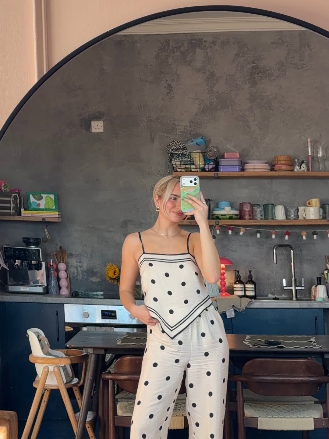 Woman in white polka dot jumpsuit taking mirror selfie in stylish kitchen with dark shelving.