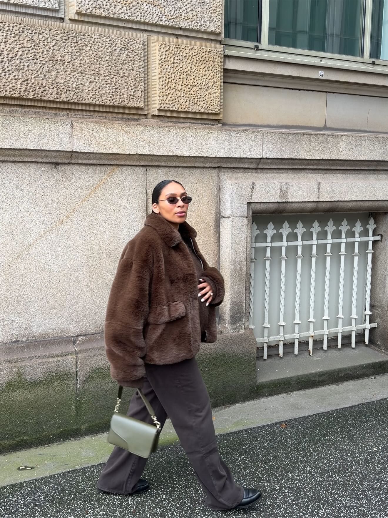 Woman in brown faux fur coat, gray trousers, and sunglasses carrying a green bag while walking on a city street.