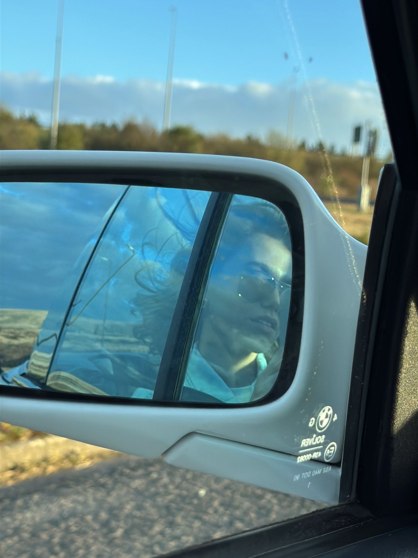 Person reflected in a BMW side mirror while driving on a highway under a blue sky.