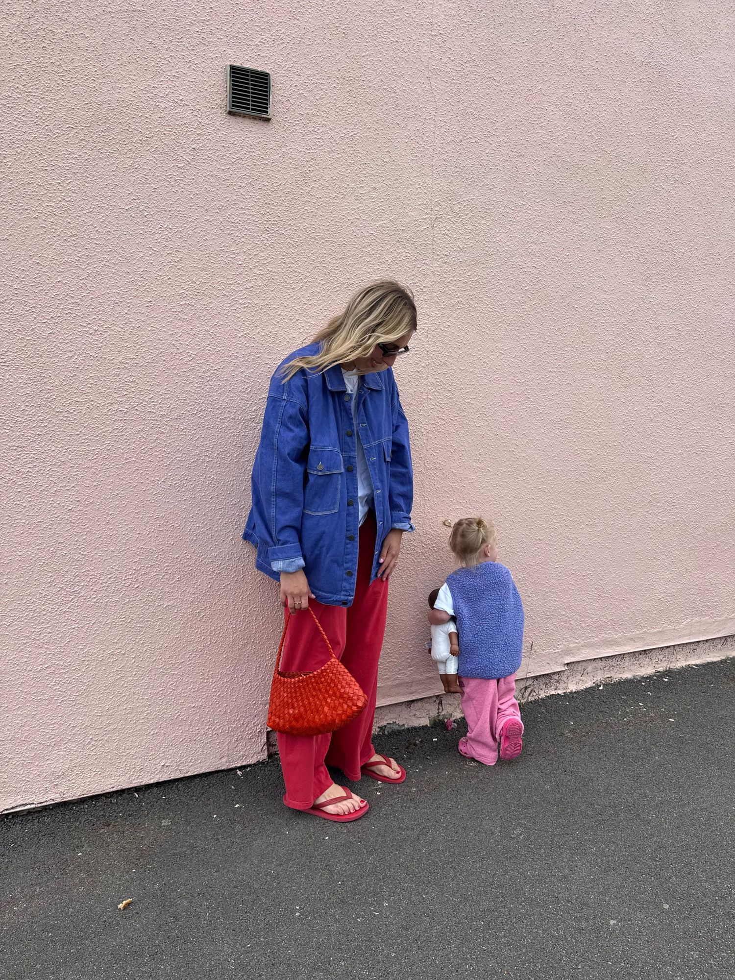 Woman in blue denim jacket and red wide-leg pants holds red bag beside toddler in pink outfit against pink wall.