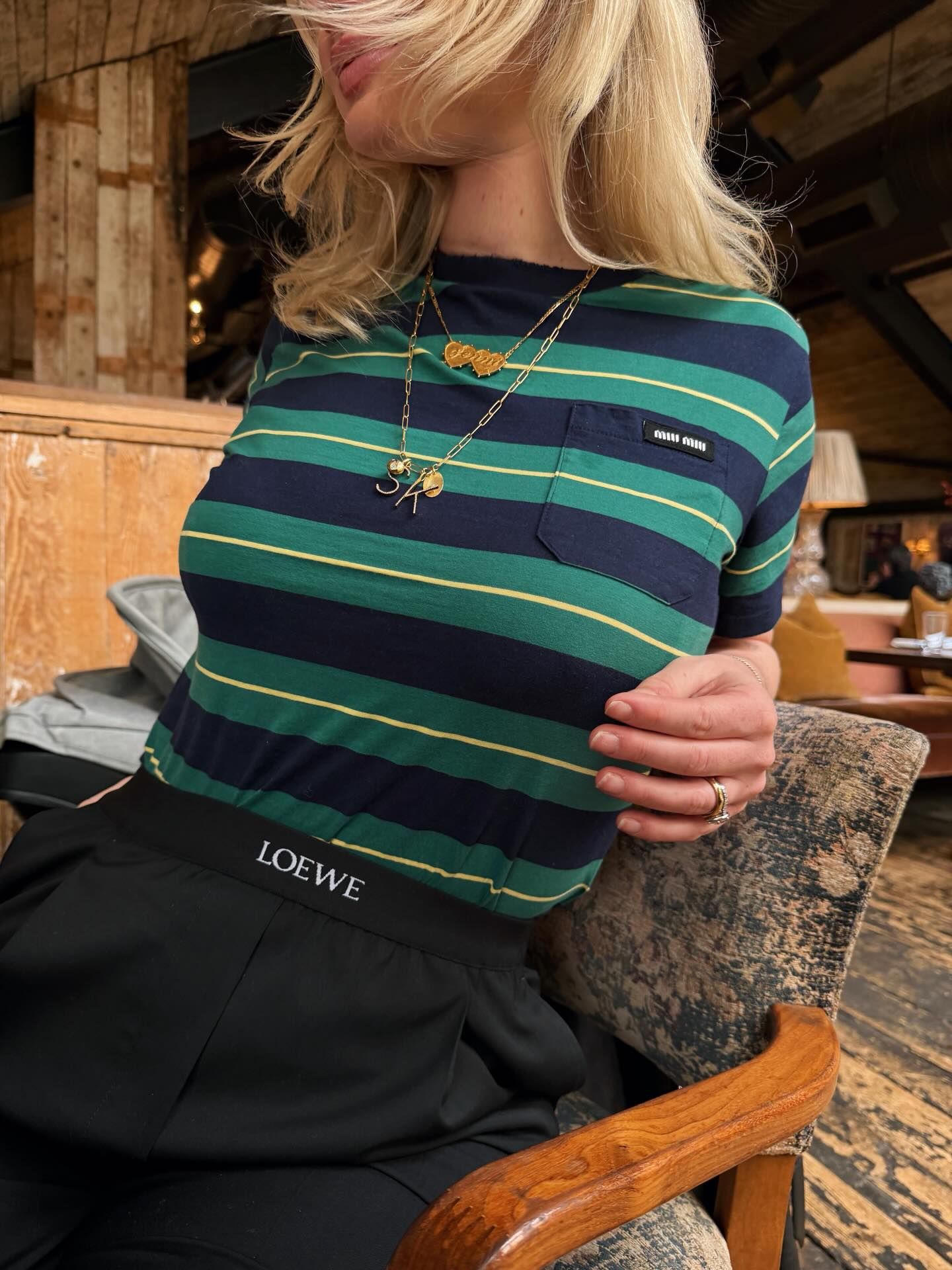 Woman in striped green navy top with gold necklace wearing Loewe black trousers seated in a restaurant.