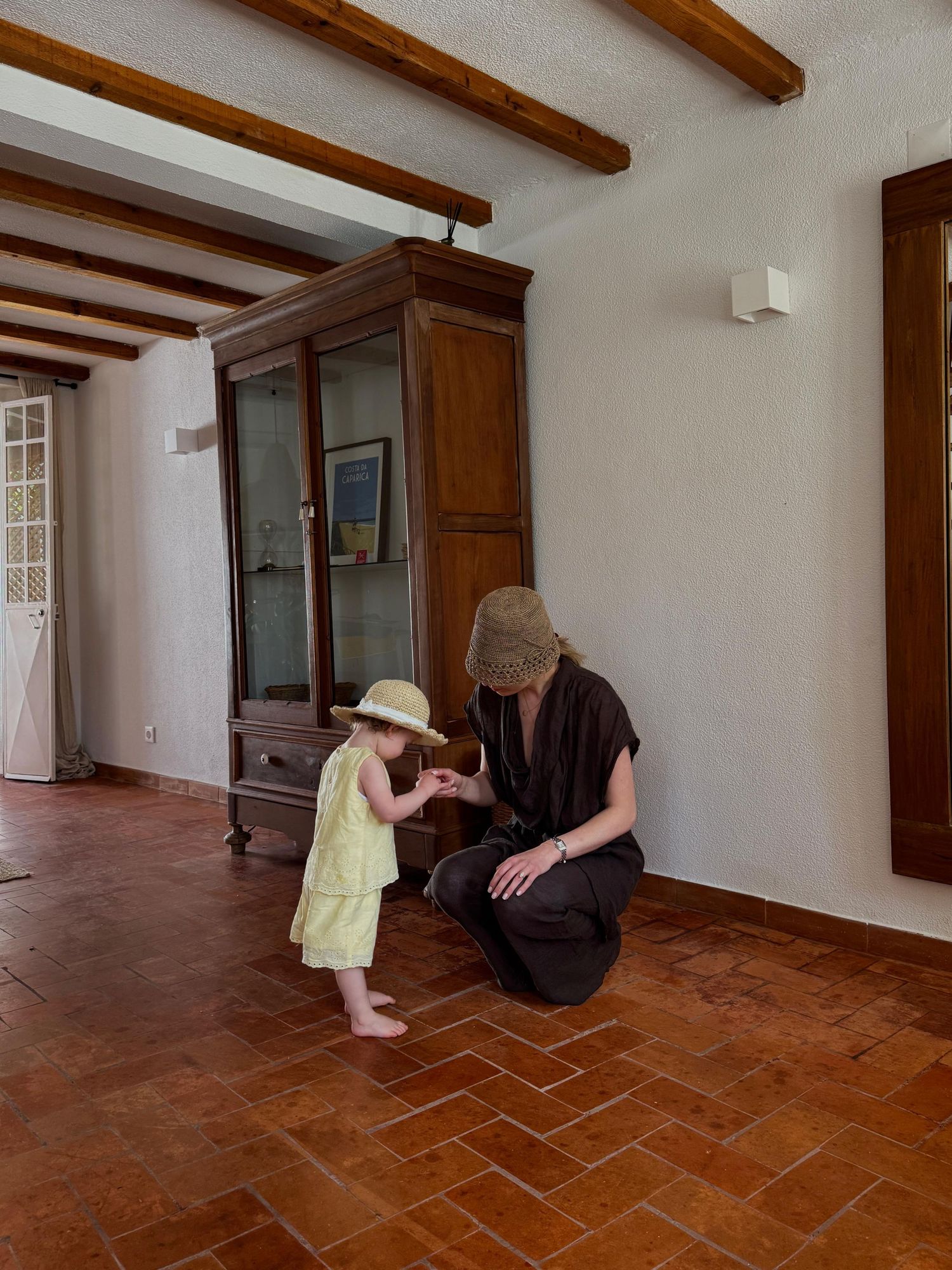 Adult crouching to interact with a toddler in a straw hat inside a rustic room with terracotta floors.
