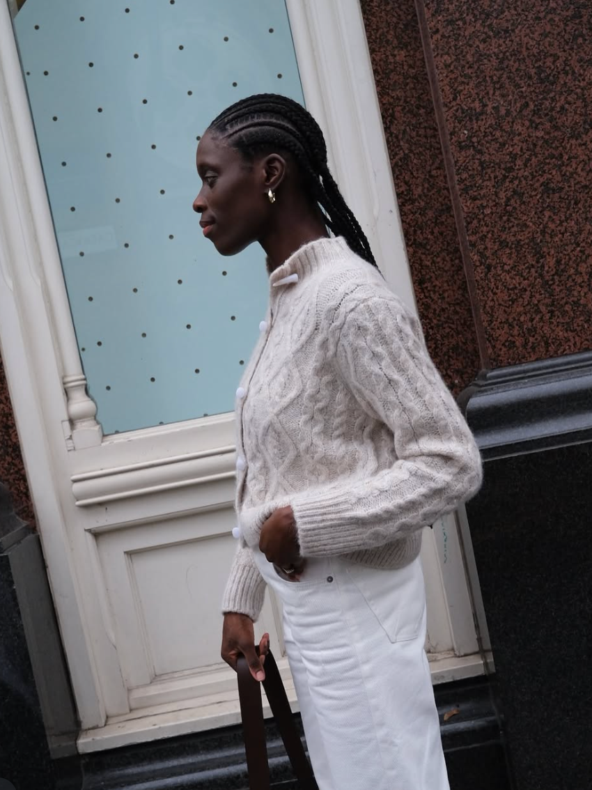 A person wearing a white cable knit sweater and white skirt stands in profile against a blue door and brown wall.