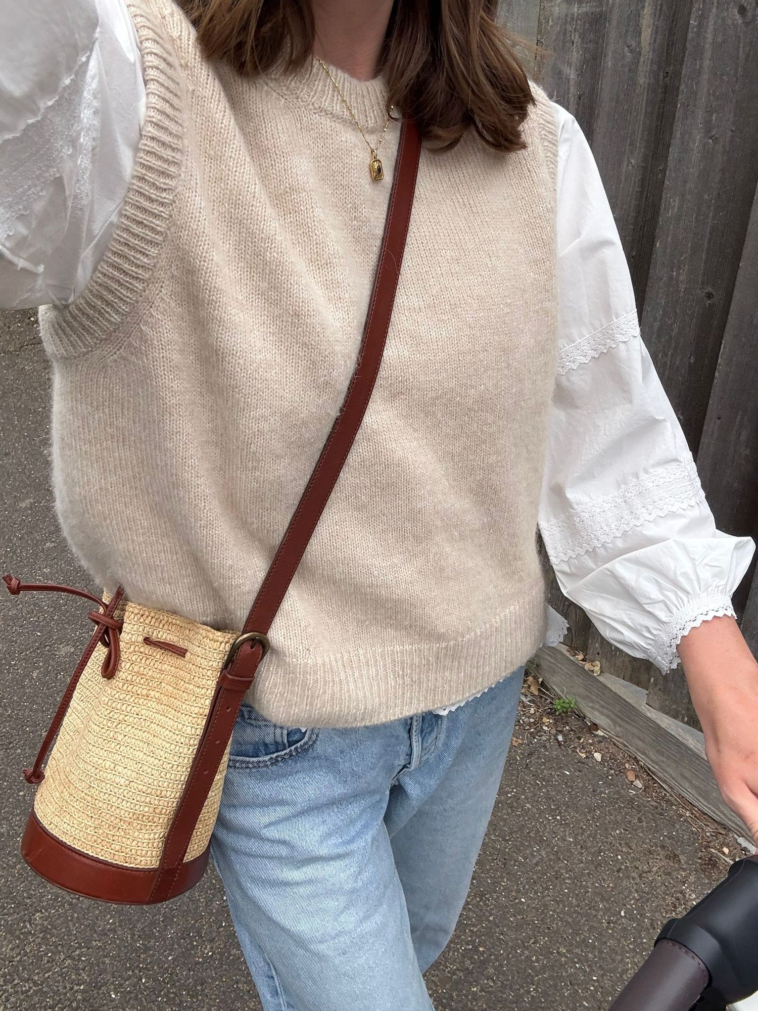 A casual outfit featuring a beige sweater vest, white blouse, blue jeans and a woven straw crossbody bag.