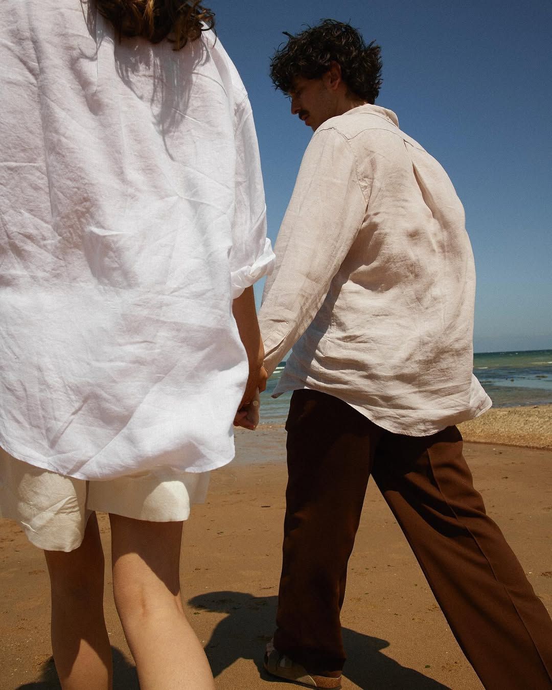 Couple holding hands walking on sunny beach, wearing linen shirts and casual summer outfits.
