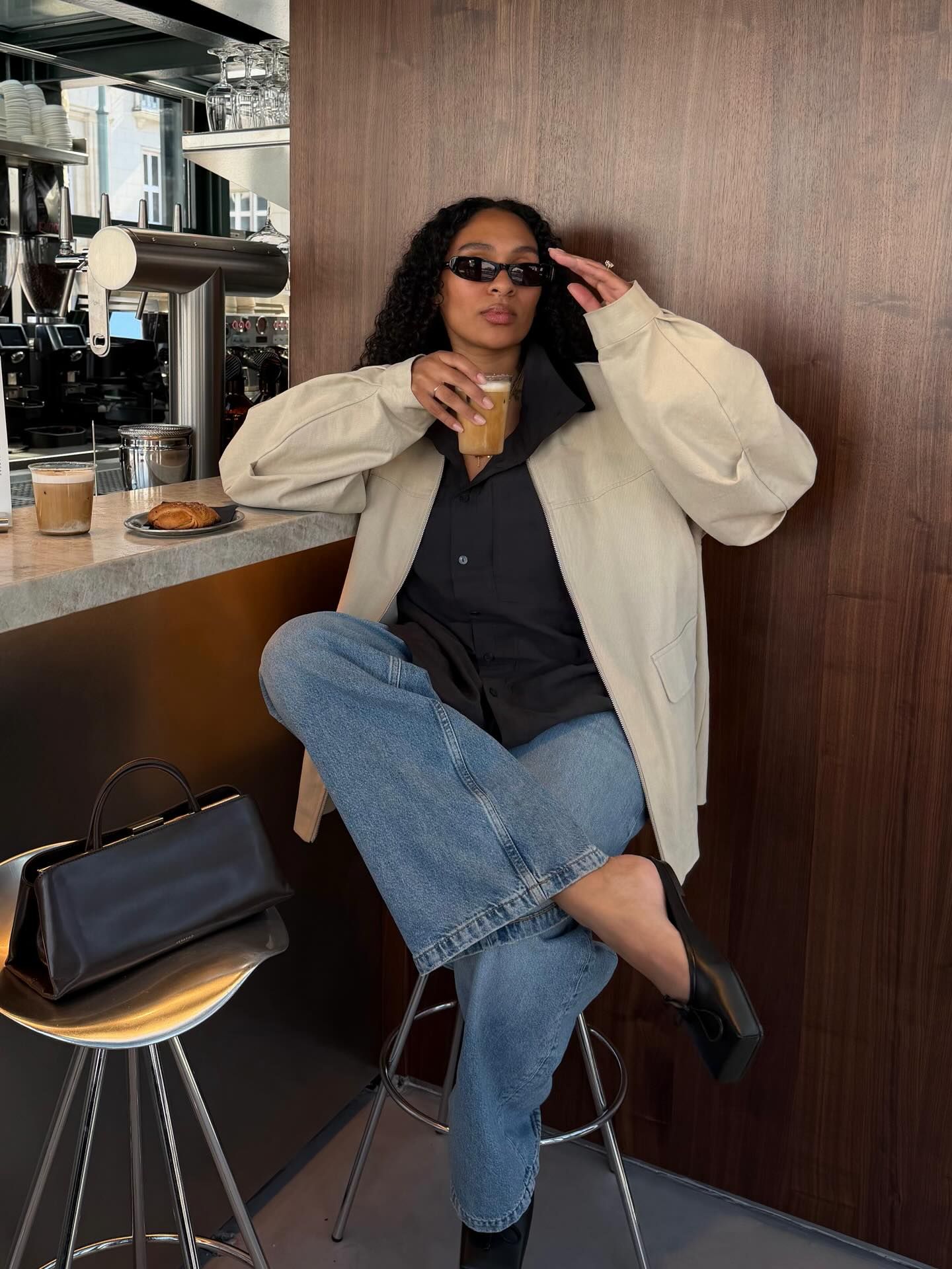 Woman in beige jacket, black shirt, jeans, and sunglasses sitting at a coffee bar holding an iced drink.