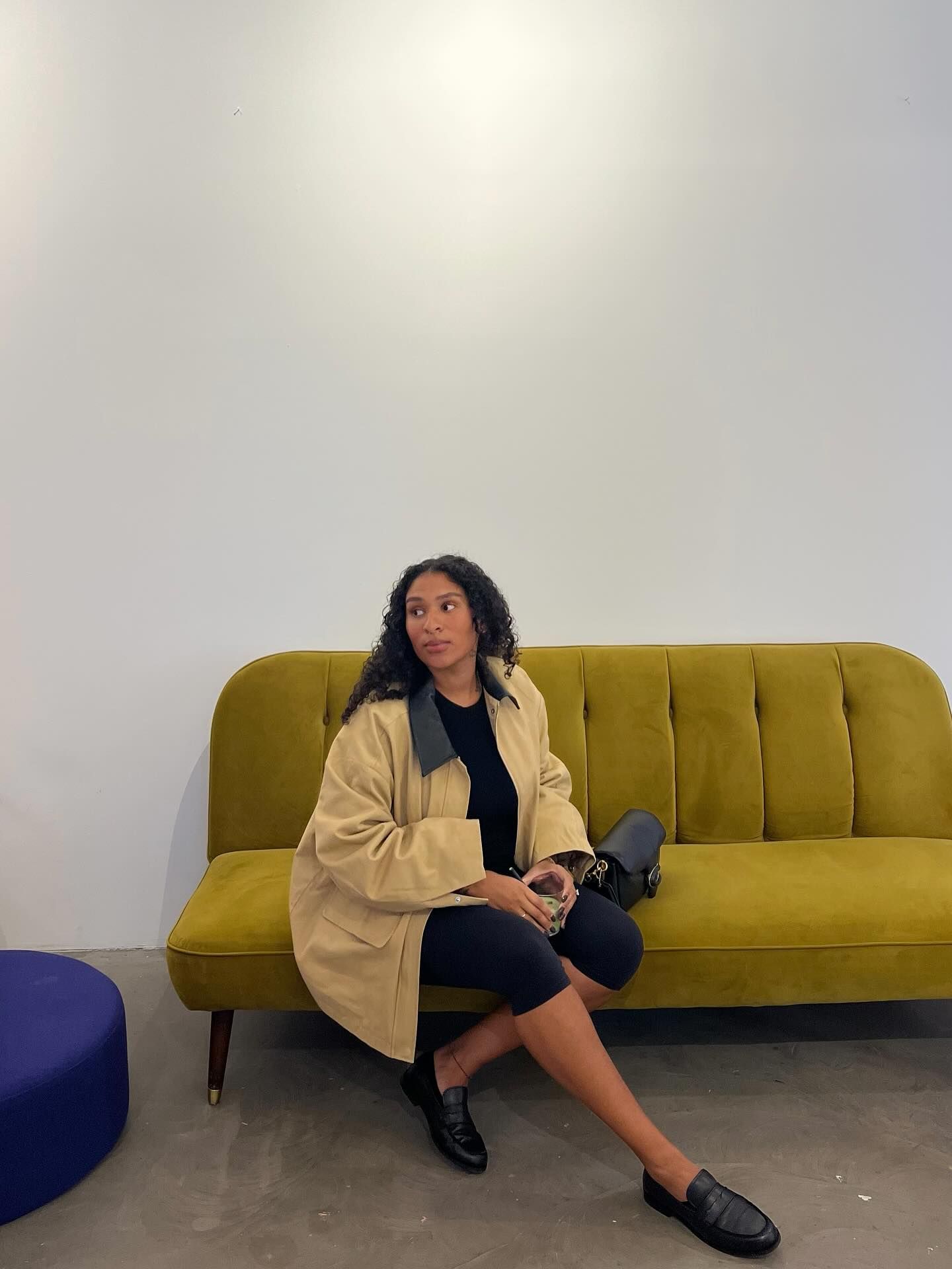 Woman in beige coat and black outfit sits on a mustard yellow velvet sofa in a minimalist white interior.