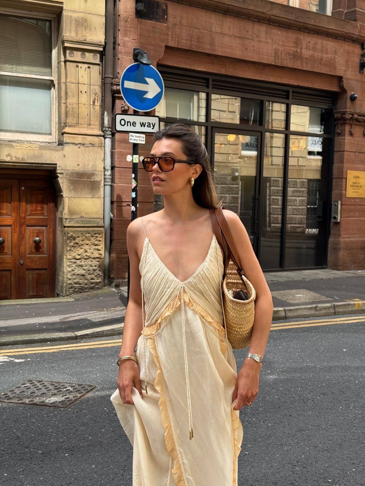 Woman in cream slip dress with woven bag and sunglasses walking on a one-way street.
