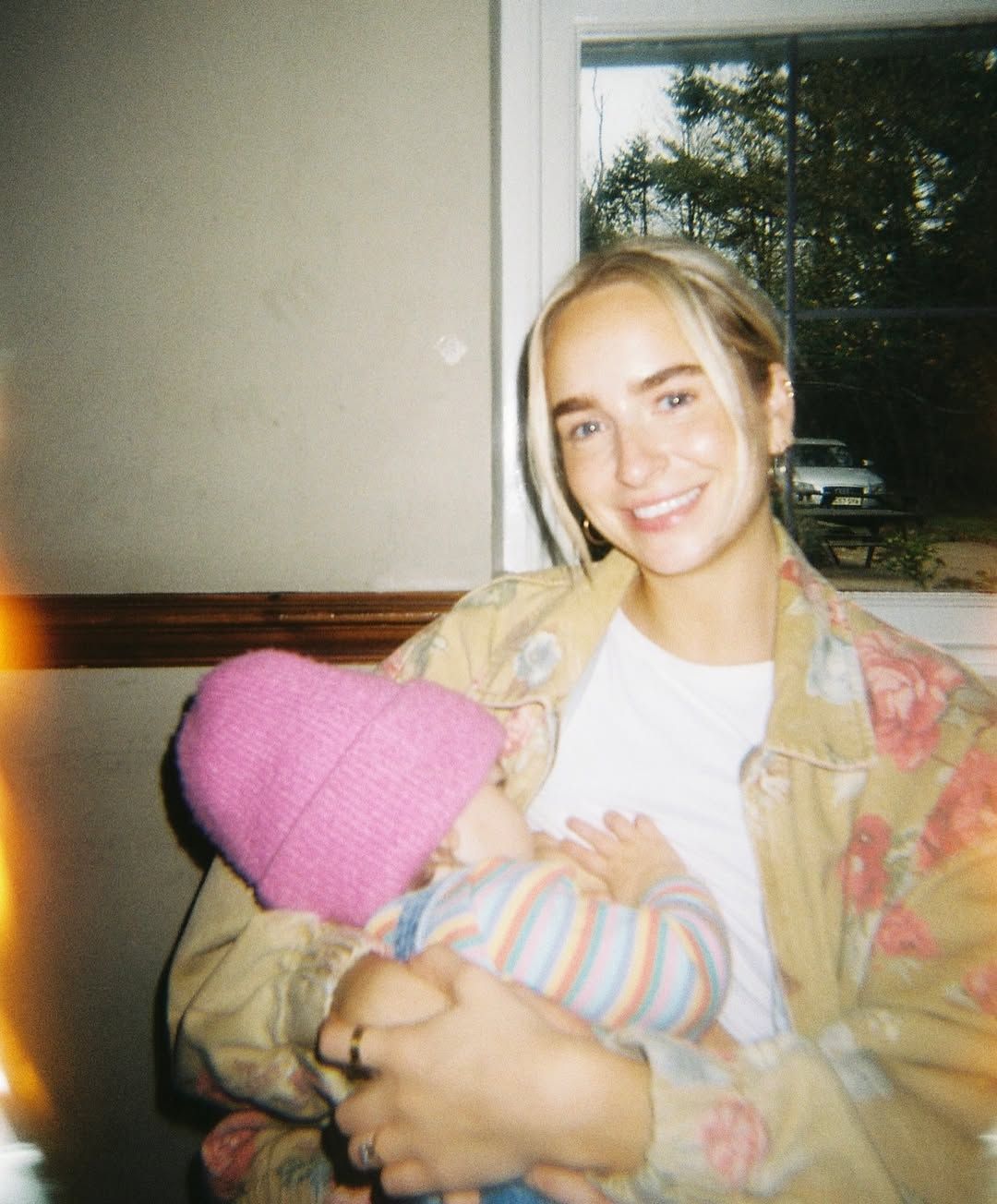Young blonde woman smiling while holding a baby in a pink knit hat indoors.