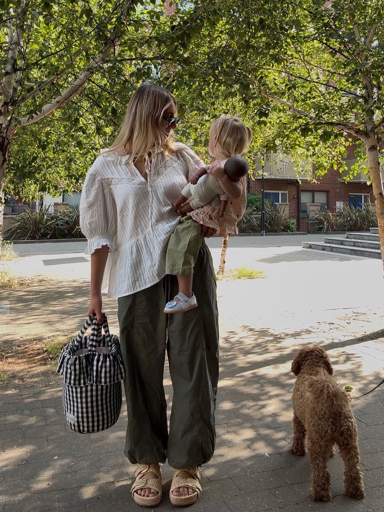 Woman in white blouse and olive trousers holds toddler while walking a brown doodle dog in a sunny park.