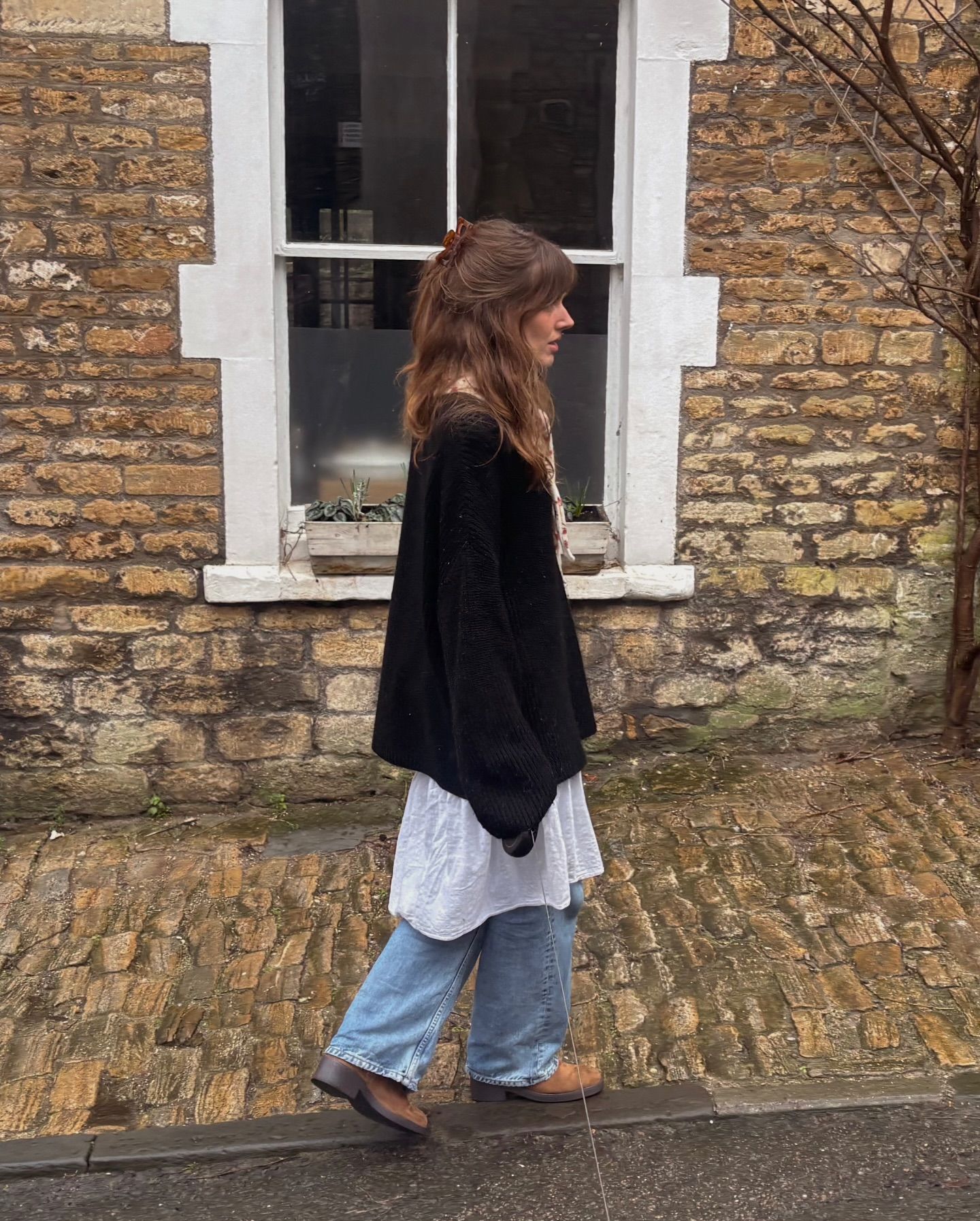 Woman in black oversized sweater, white shirt, blue jeans, and brown flats walking on cobblestone street by brick building.