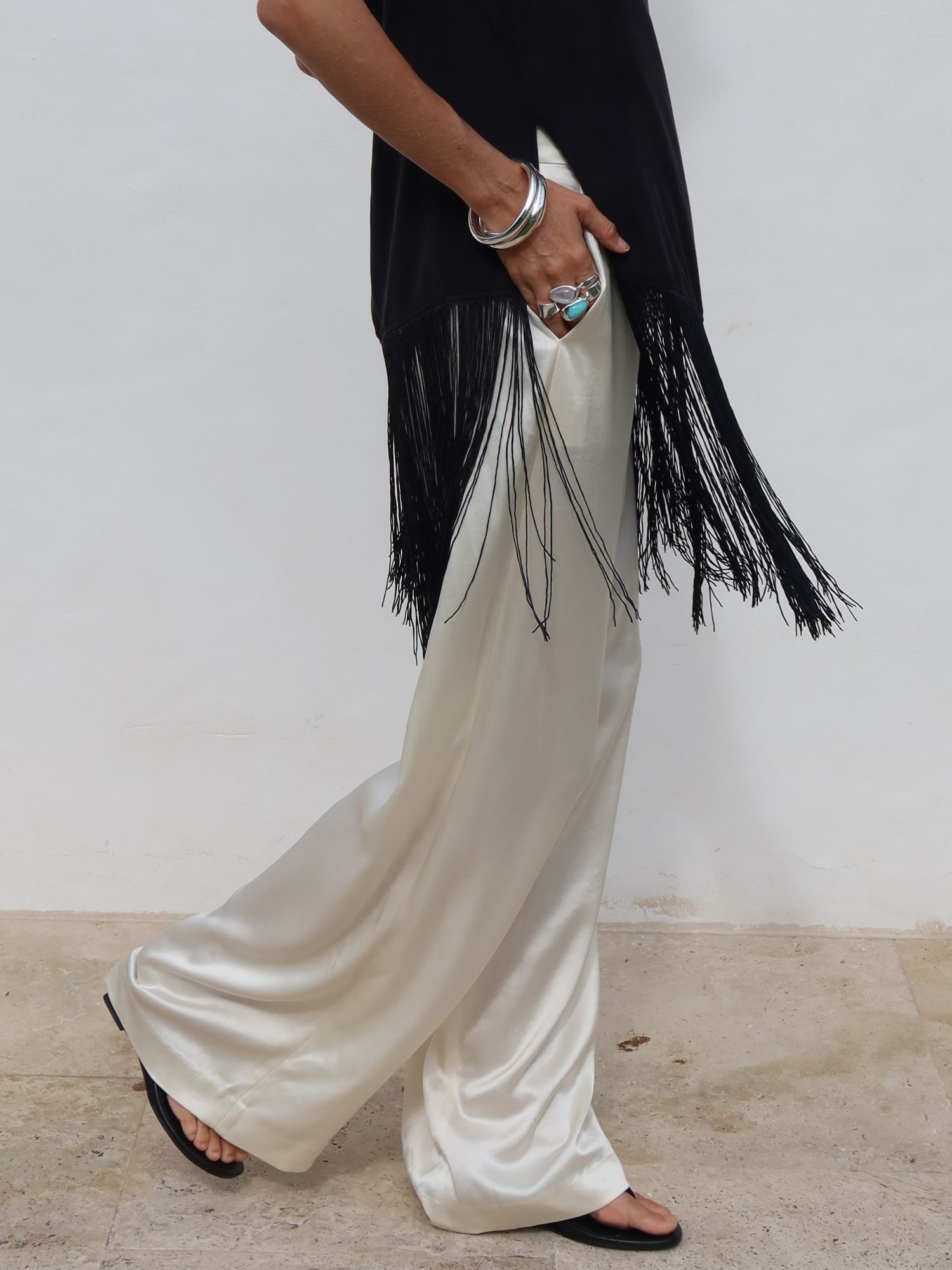 Model wearing black fringe top with ivory satin wide-leg pants and black sandals, styled with silver bracelets.