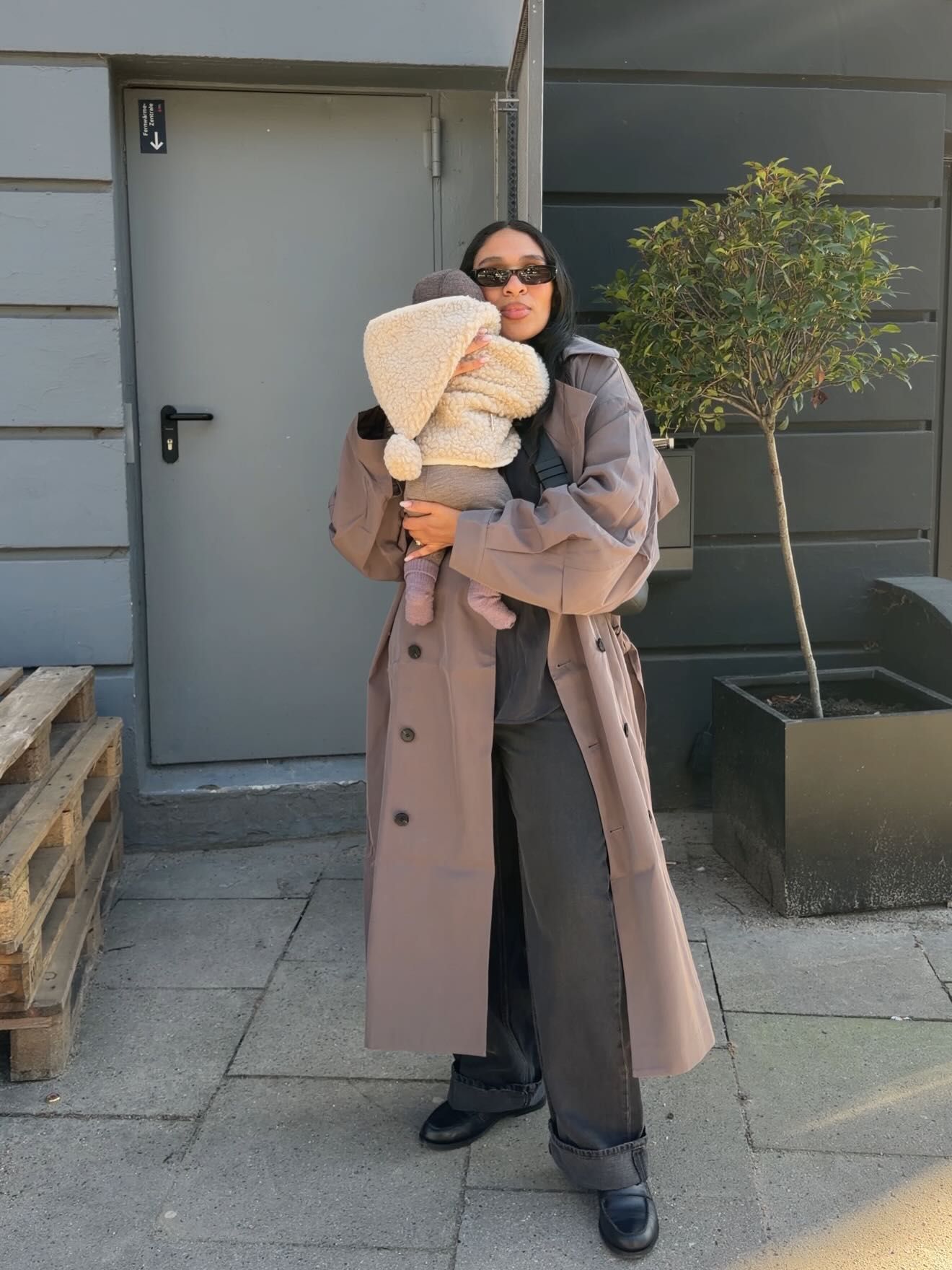 Woman in taupe trench coat holding a baby outdoors.