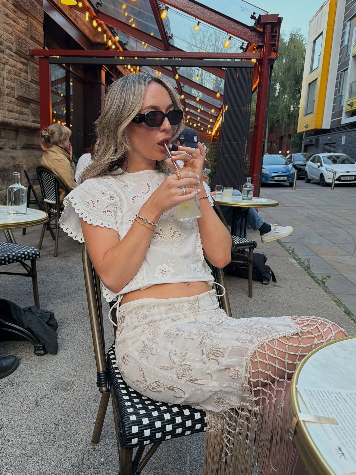 Woman in white lace crop top and skirt sipping iced drink at outdoor cafe patio wearing sunglasses.