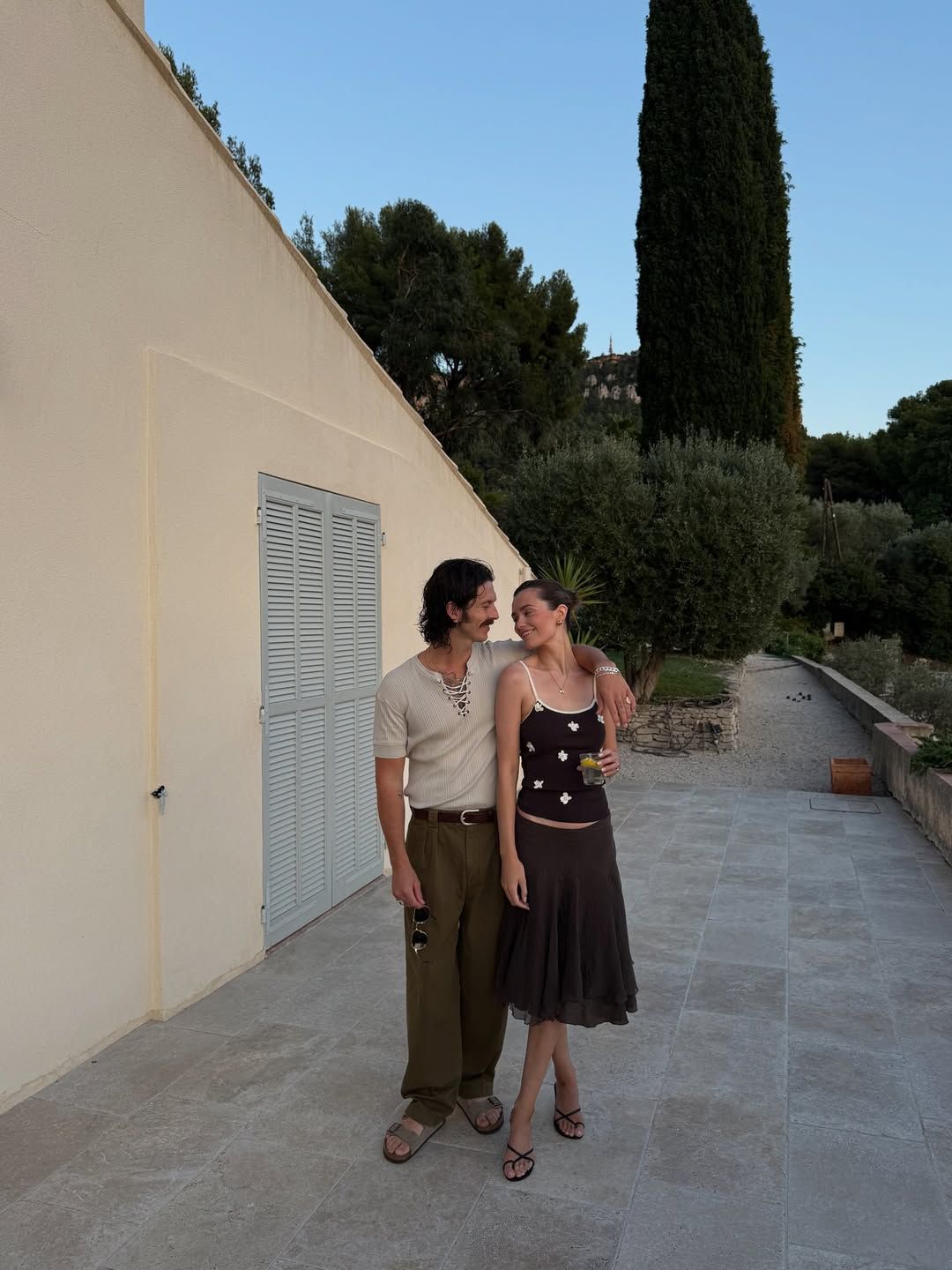 Couple walking together on a stone terrace beside a white villa with cypress trees at dusk.