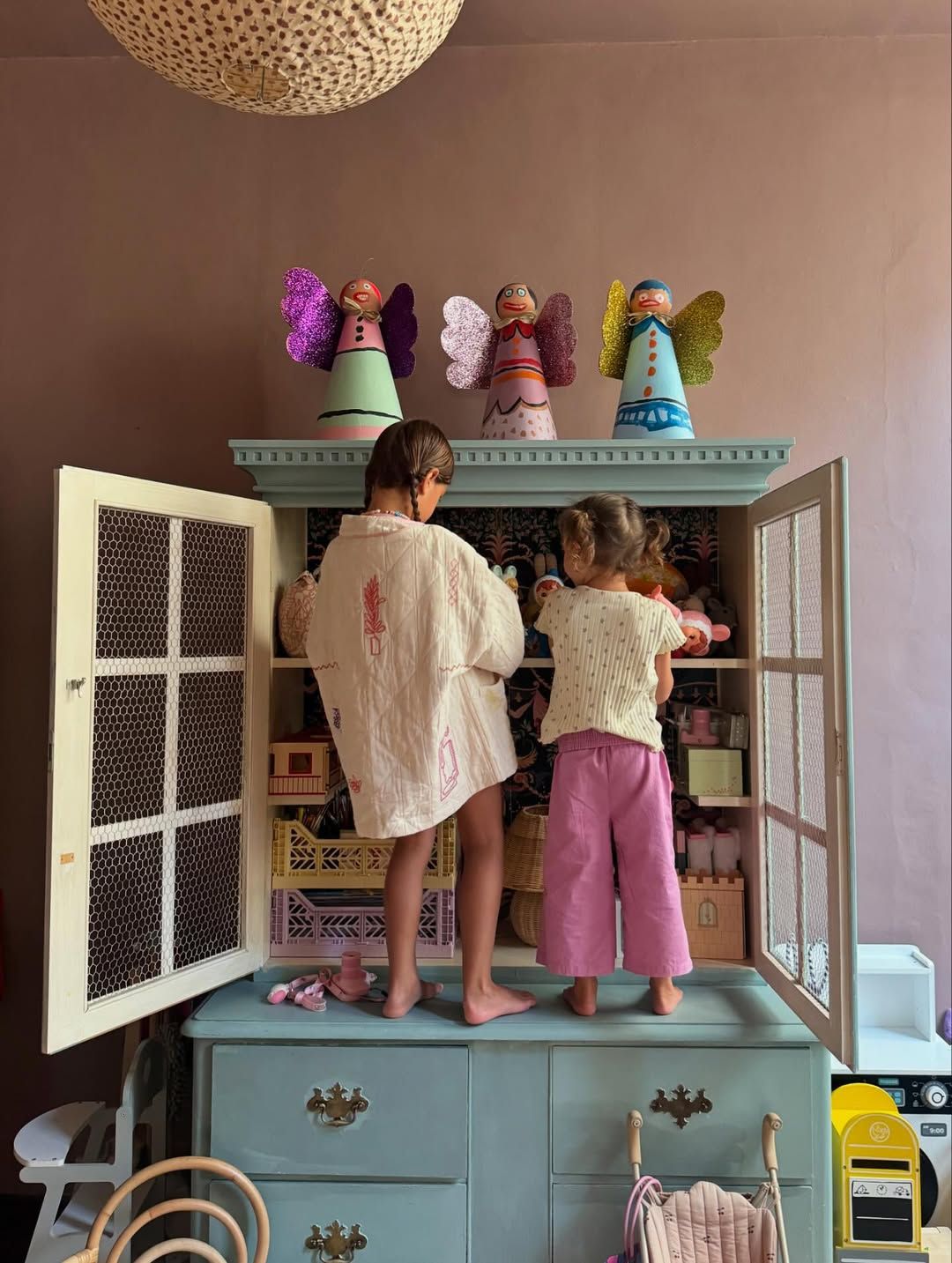 Two young girls stand on a teal dresser exploring a dollhouse cabinet with angel figurines on top.