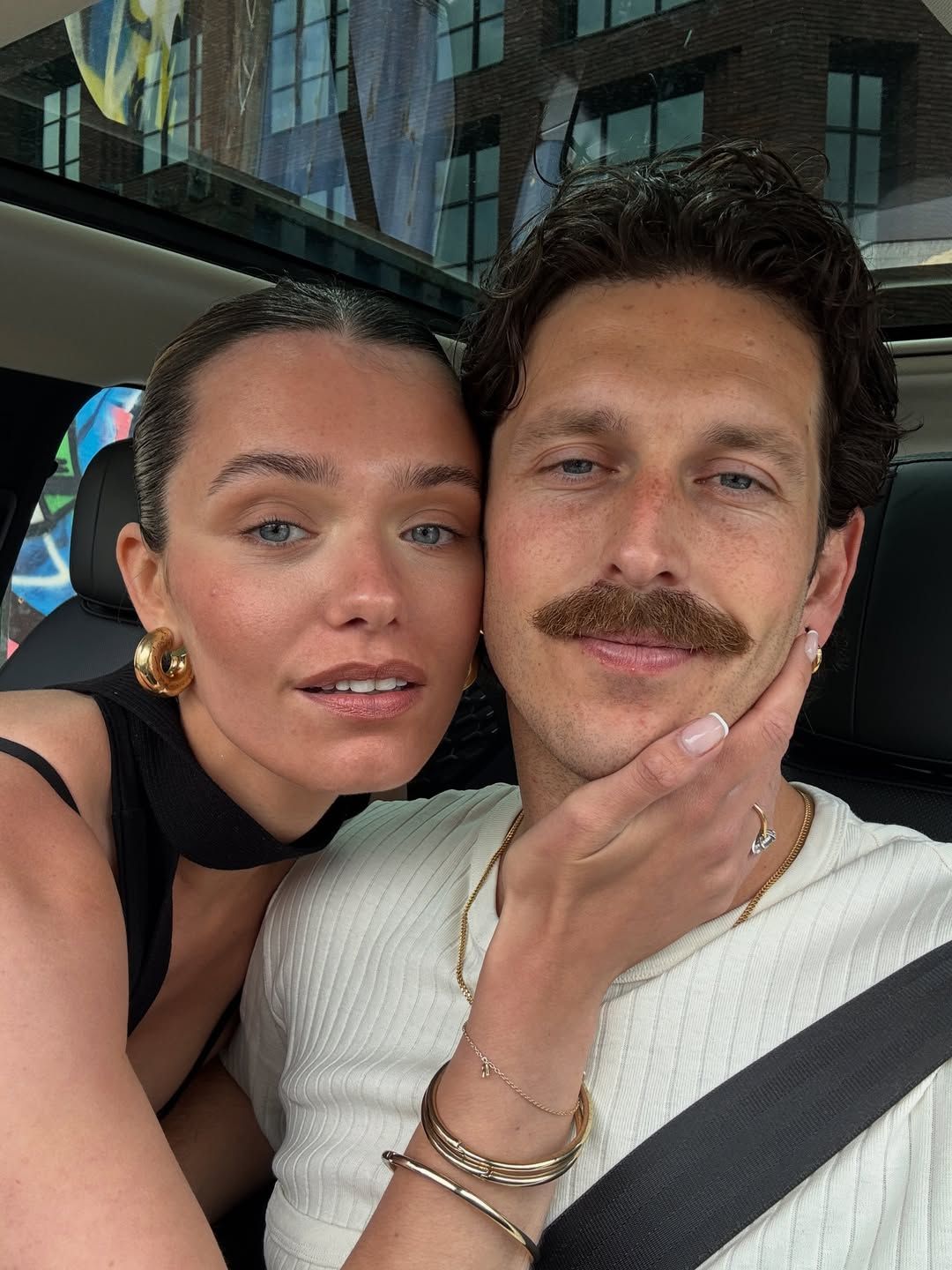 A couple takes a selfie in a car, woman in black top with gold earrings leans on man with mustache in white shirt.