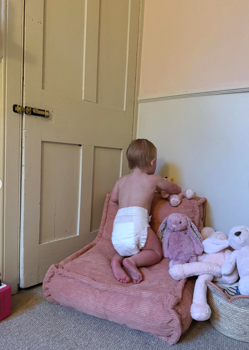 A baby in a diaper crawls on a pink cushion with stuffed animals and toys in a bright room.