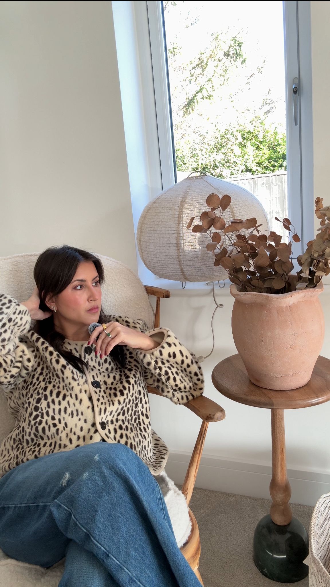 A person in a leopard print sweater sits near a decorative hat stand displaying mushroom-adorned hats in a bright room.