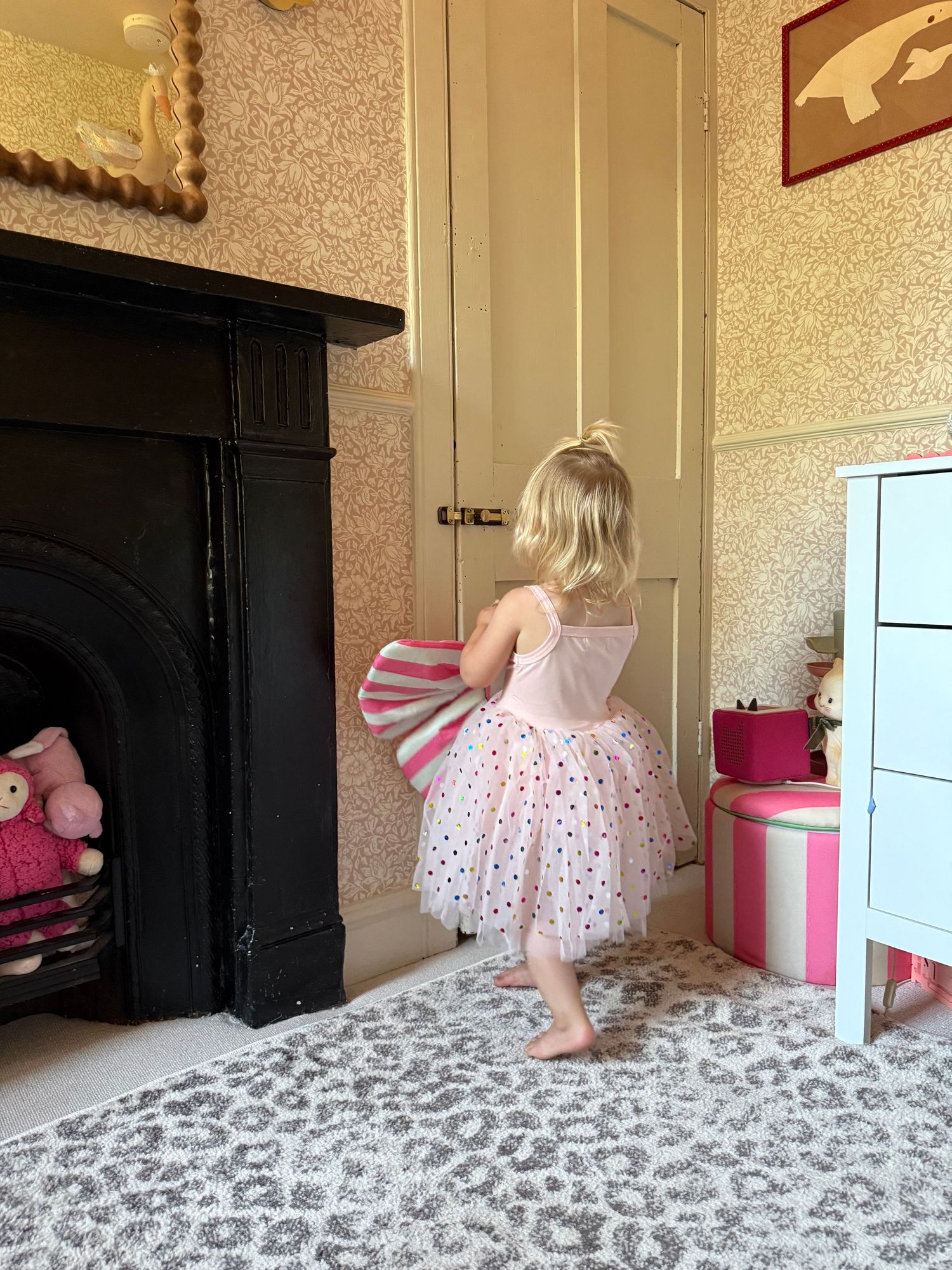A young girl in a pink tutu dress holds a striped bag near a Victorian fireplace in a decorated children's bedroom.