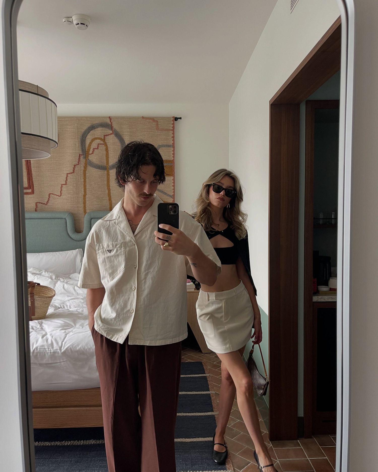 Couple taking a mirror selfie in a hotel room, man in white shirt and brown pants, woman in black crop top and mini skirt.