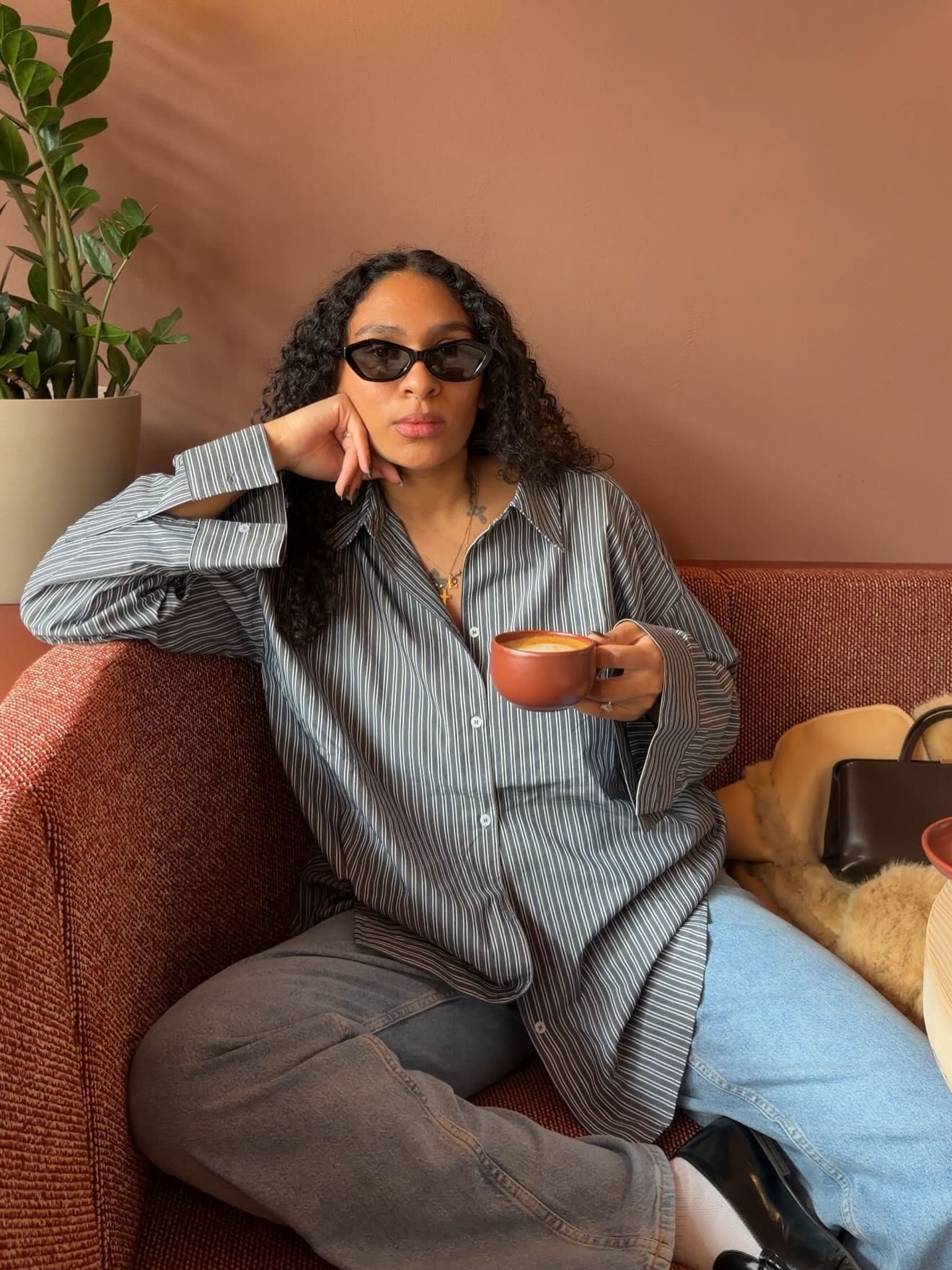 Woman in striped oversized shirt and jeans holding a coffee cup, wearing sunglasses, relaxing on a couch.