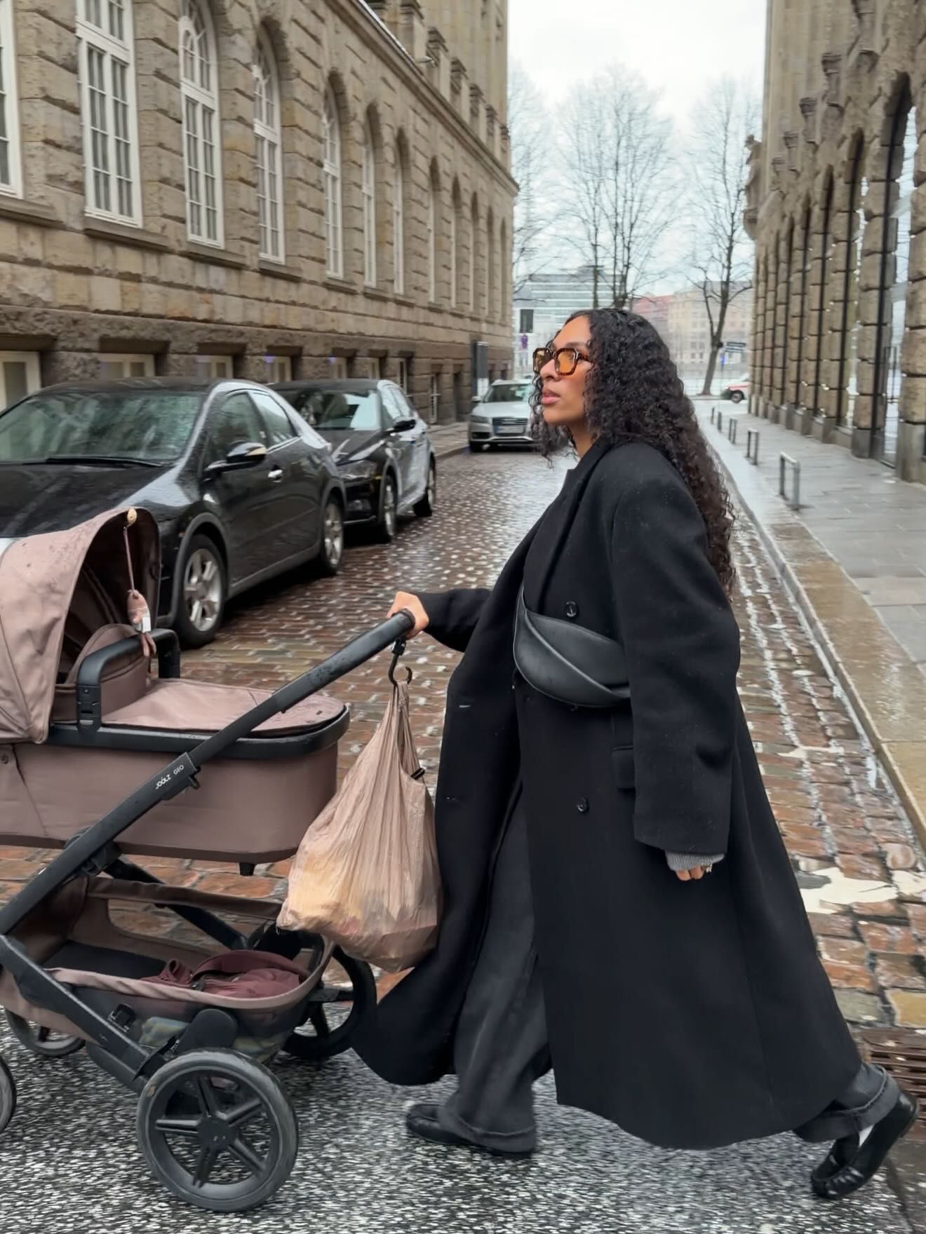 Woman in black coat pushing a baby stroller on a cobblestone street in a European city.