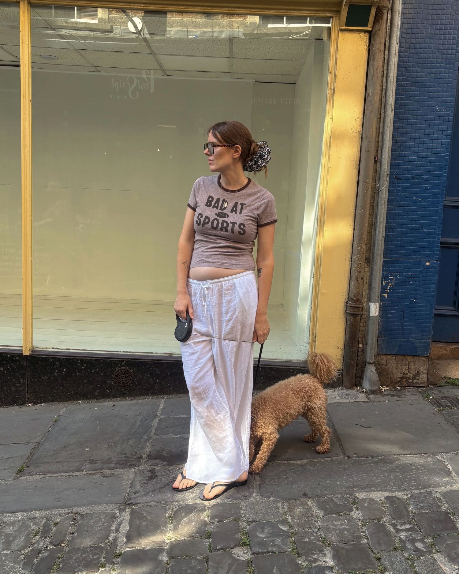Woman in 'Bad at Sports' tee and white maxi skirt walks a golden doodle on a cobblestone street.