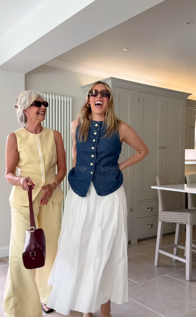 Two women laughing in stylish outfits, one in yellow dress, other in denim vest and white skirt, standing in modern kitchen.