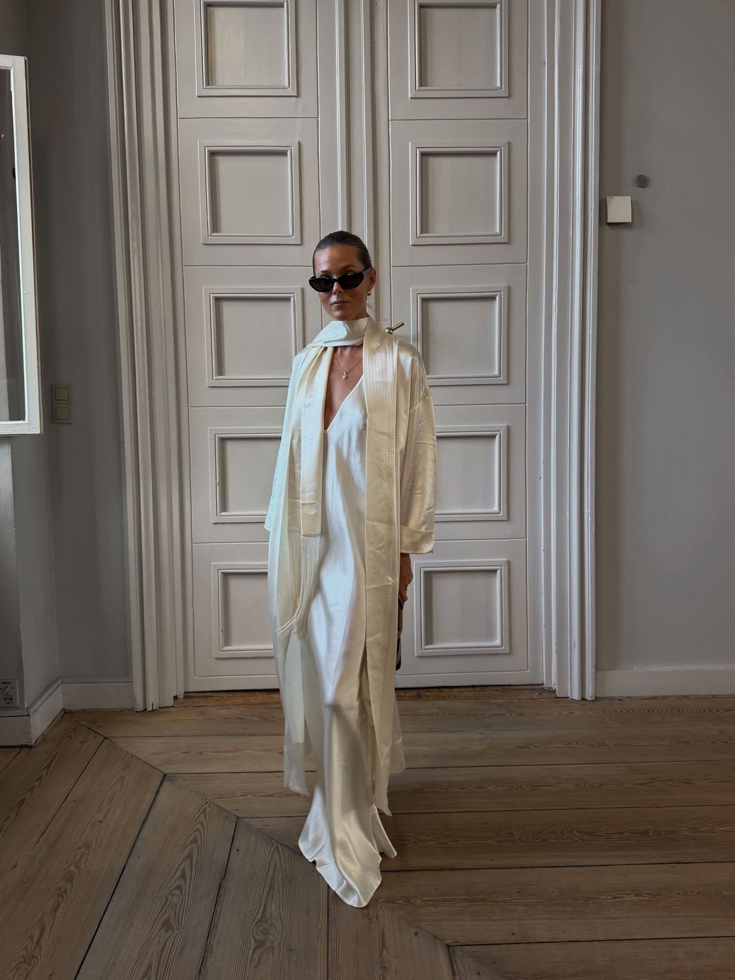 Woman in cream silk slip dress and long ivory coat with sunglasses standing before white paneled doors.