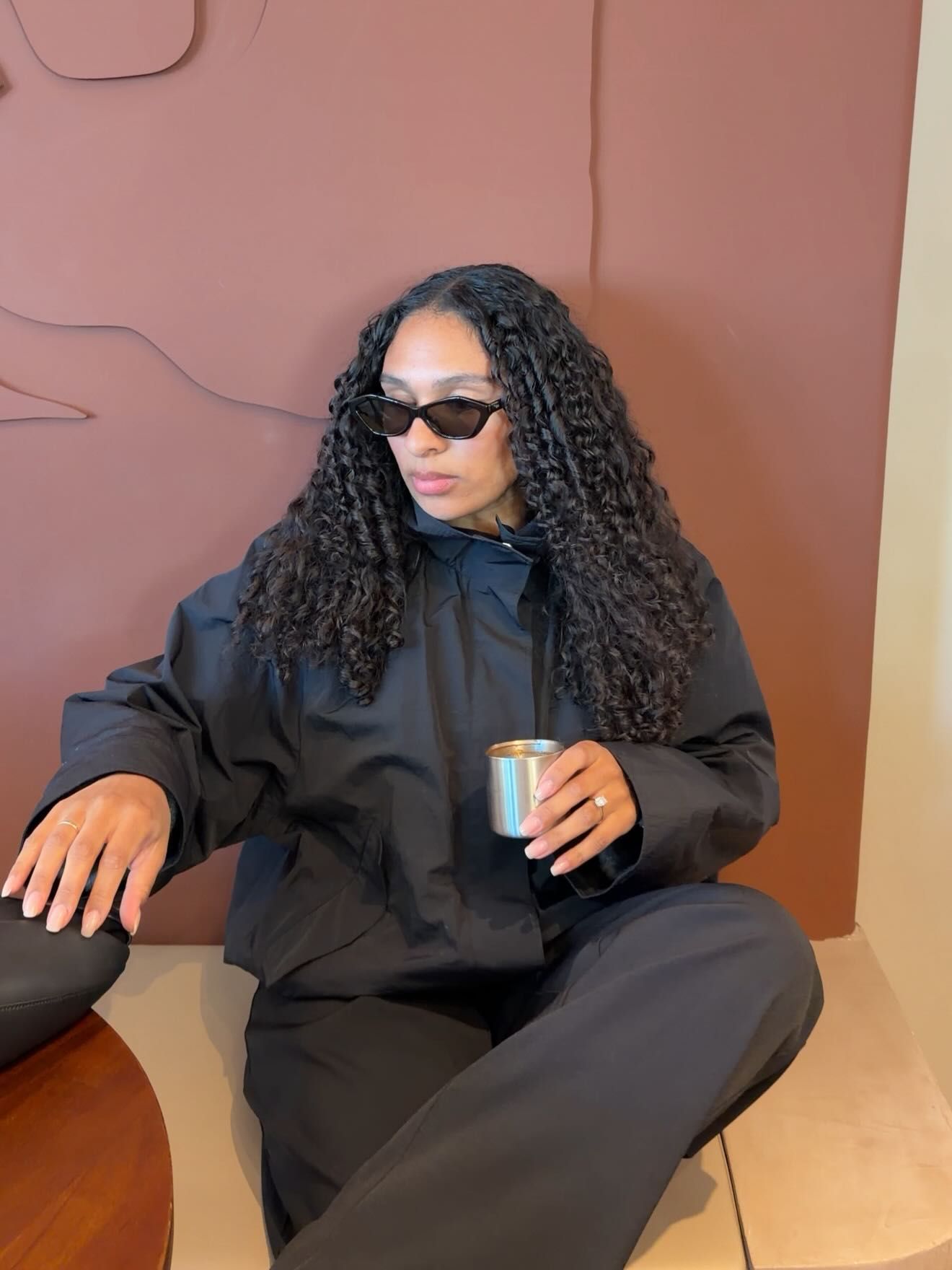A woman with long curly black hair wearing all-black outfit and sunglasses sits cross-legged holding a metallic cup.