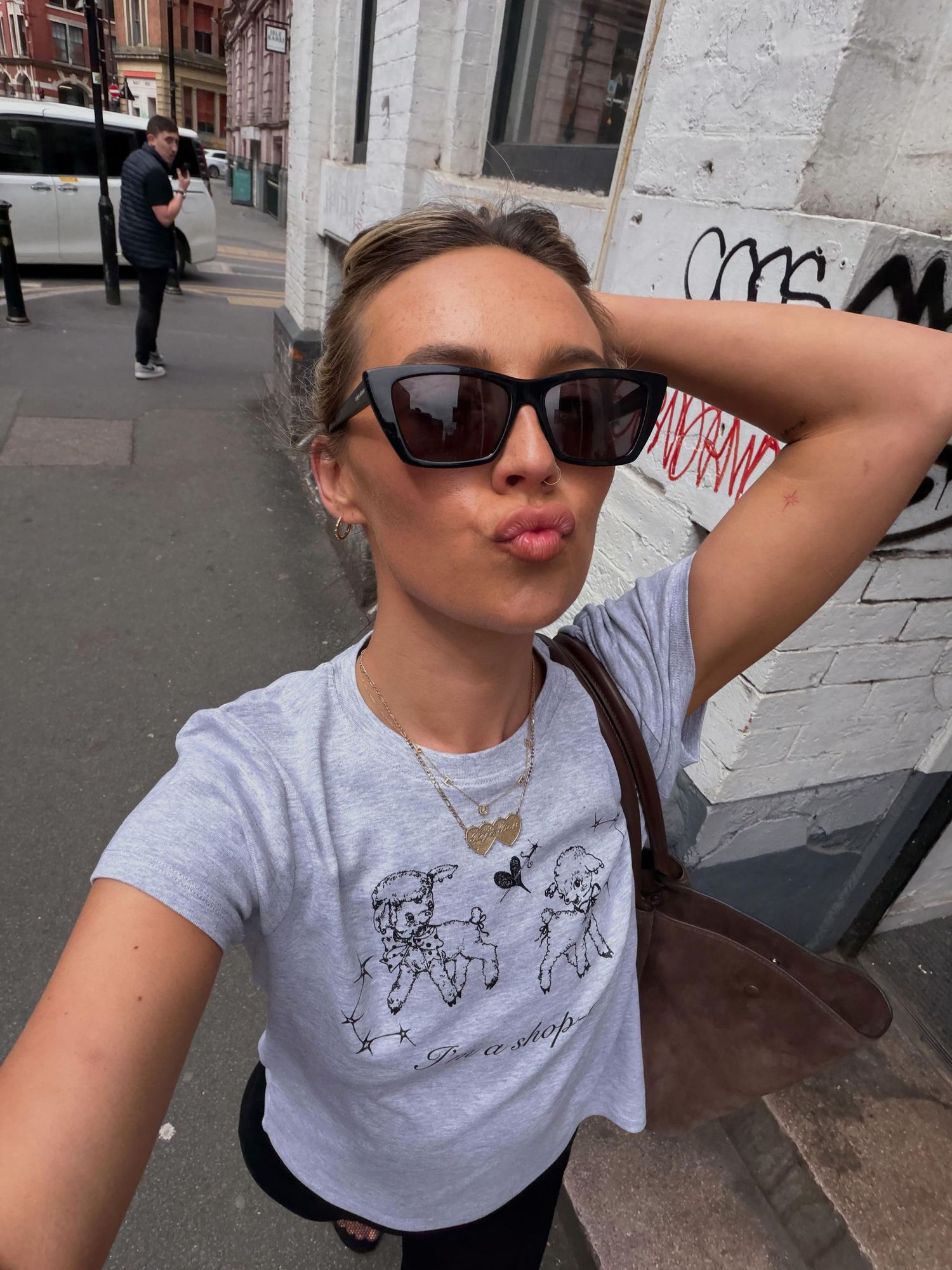 Young woman in sunglasses and graphic tee takes a selfie on a city street, posing with a duck face.