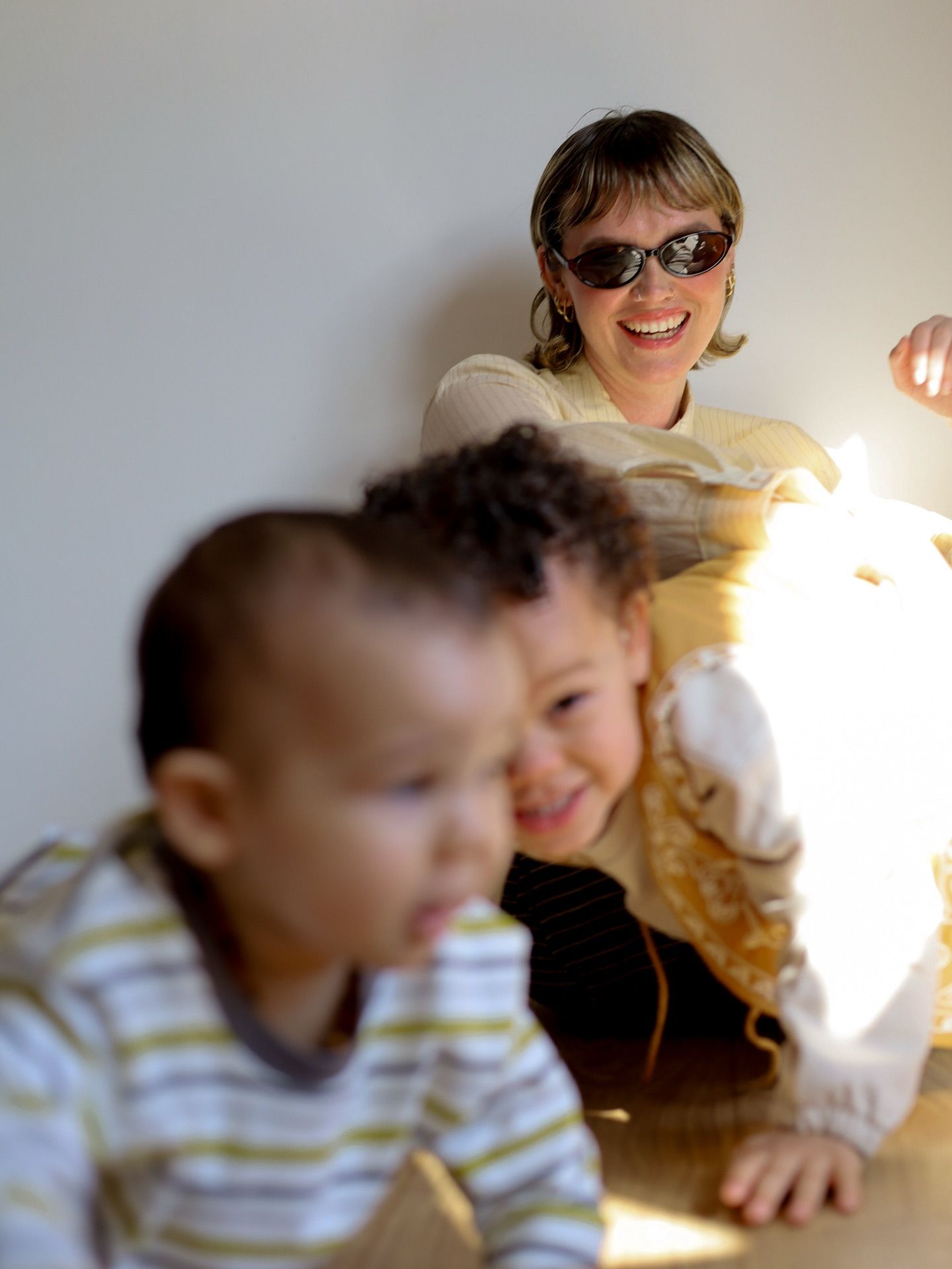 Woman in sunglasses smiling at two young children playing in warm sunlight indoors.
