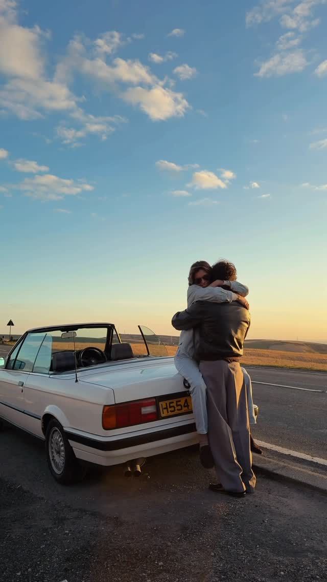 Two people embracing beside a white convertible car at sunset on an open road.